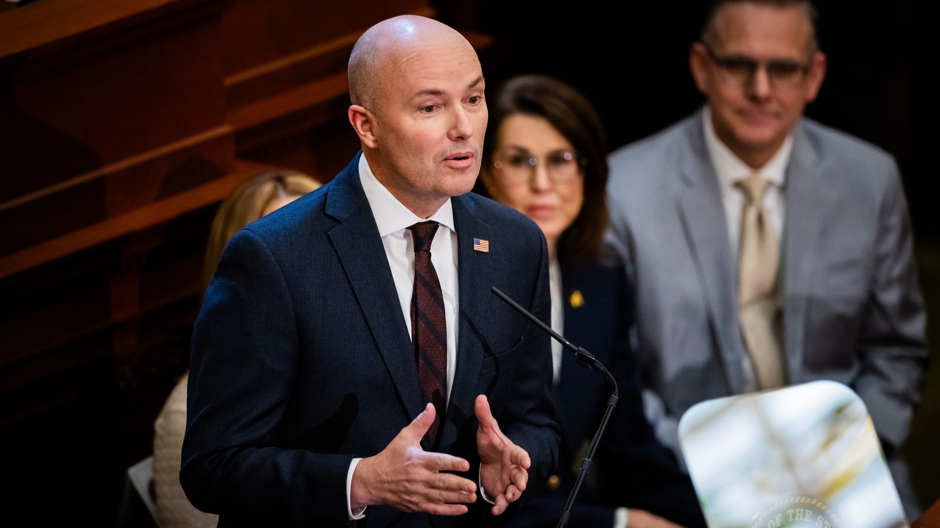 A bald man in a dark suit and maroon tie speaks at a podium with a microphone. Two people sit behind him, one woman in glasses and one man in a light gray suit and tie.