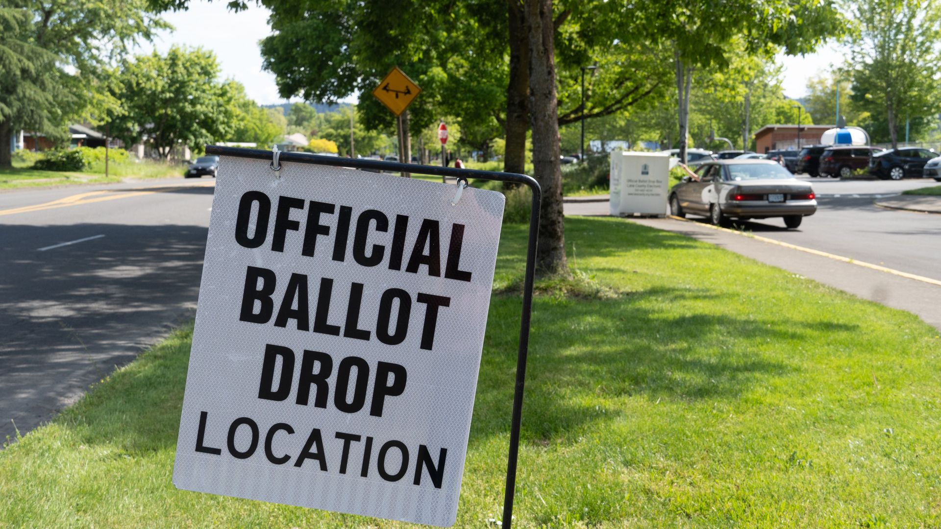An official ballot drop location sign is seen with a driver dropping off a ballot in the background.