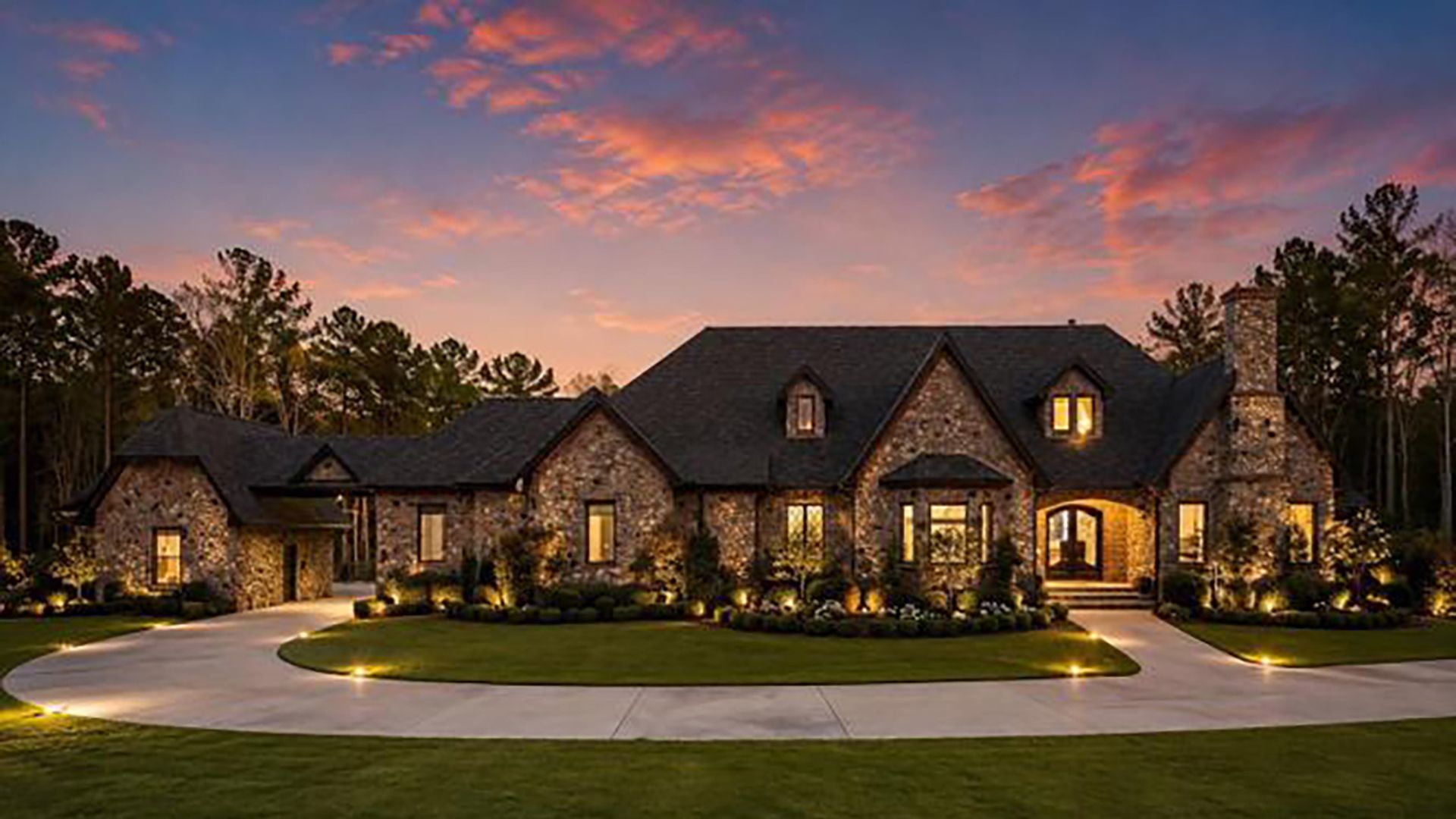 Large stone house at dusk with warm-lit windows, a curved driveway around a manicured lawn, and trees in the background; bottom-right watermark reads "For Sale".