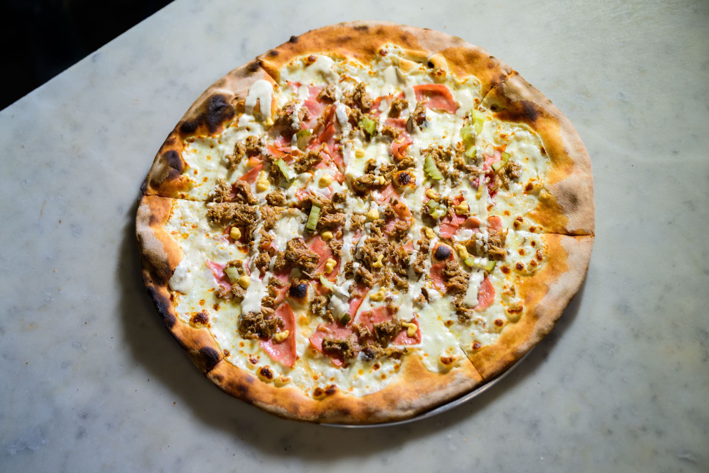 Thin-crust pizza with melted cheese, shredded meat, sliced pickles, and drizzled sauce on a marble surface.