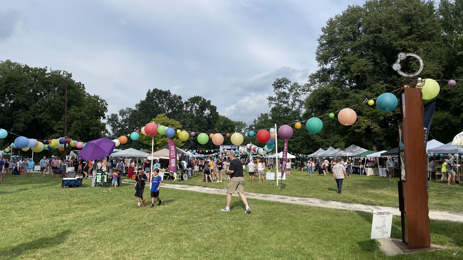 Outdoor festival with colorful paper lanterns hanging on strings, artist tents labeled "Artist Row," people walking on grass and pathways under a cloudy sky surrounded by trees.