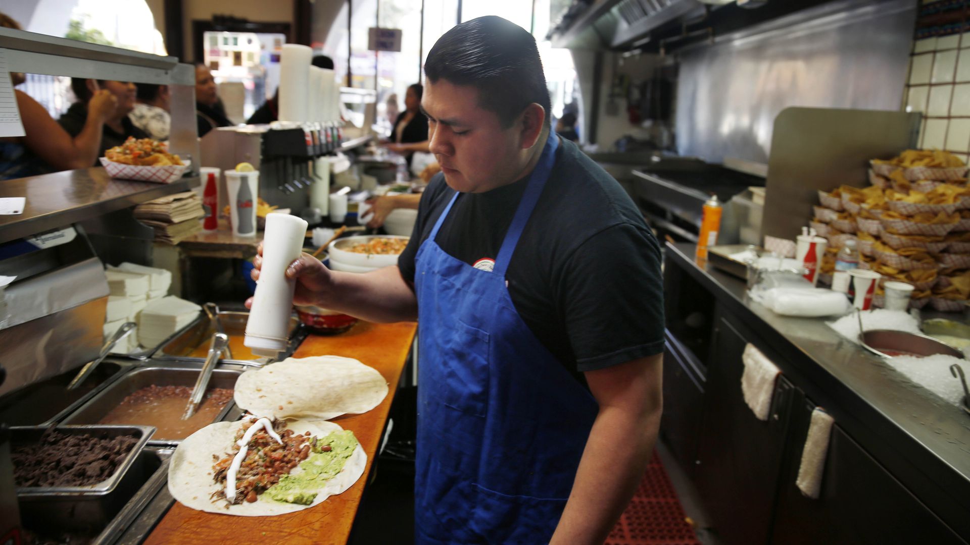 A man wearing a blue apron prepares a burrito in a taqueria.