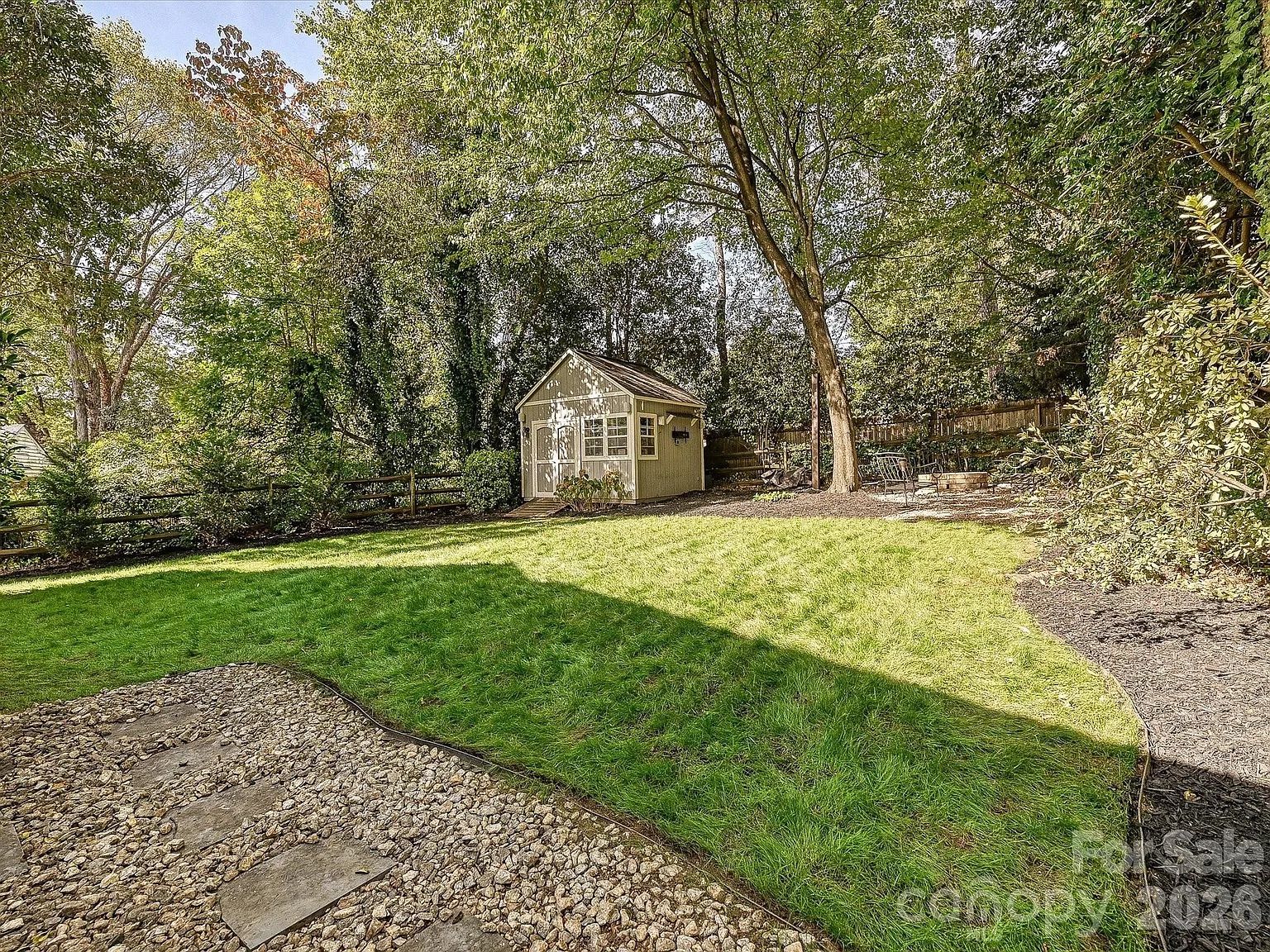 Backyard with green grass, gravel pathway with stepping stones, small light-colored shed, tall trees, and seating area around a fire pit in a fenced yard on a sunny day.