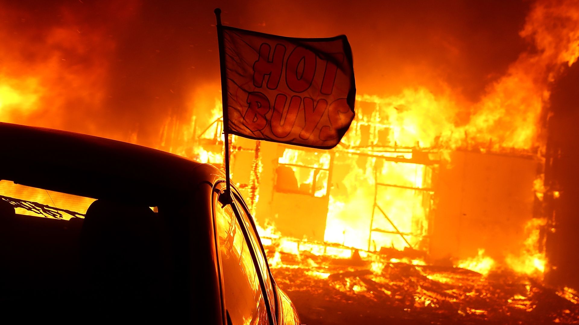 A flag that says "Hot Buys" is on a car in front of a burning car dealership