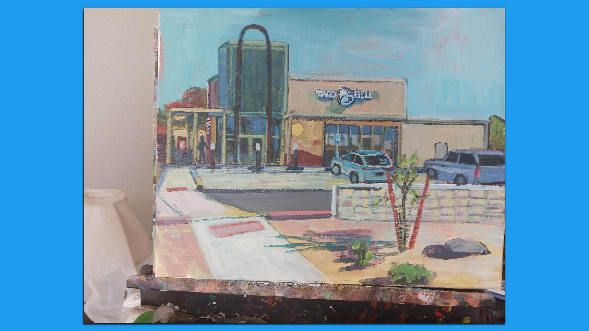 Painting of a Taco Bell restaurant with two parked cars, a sidewalk and desert landscaping in the foreground. 