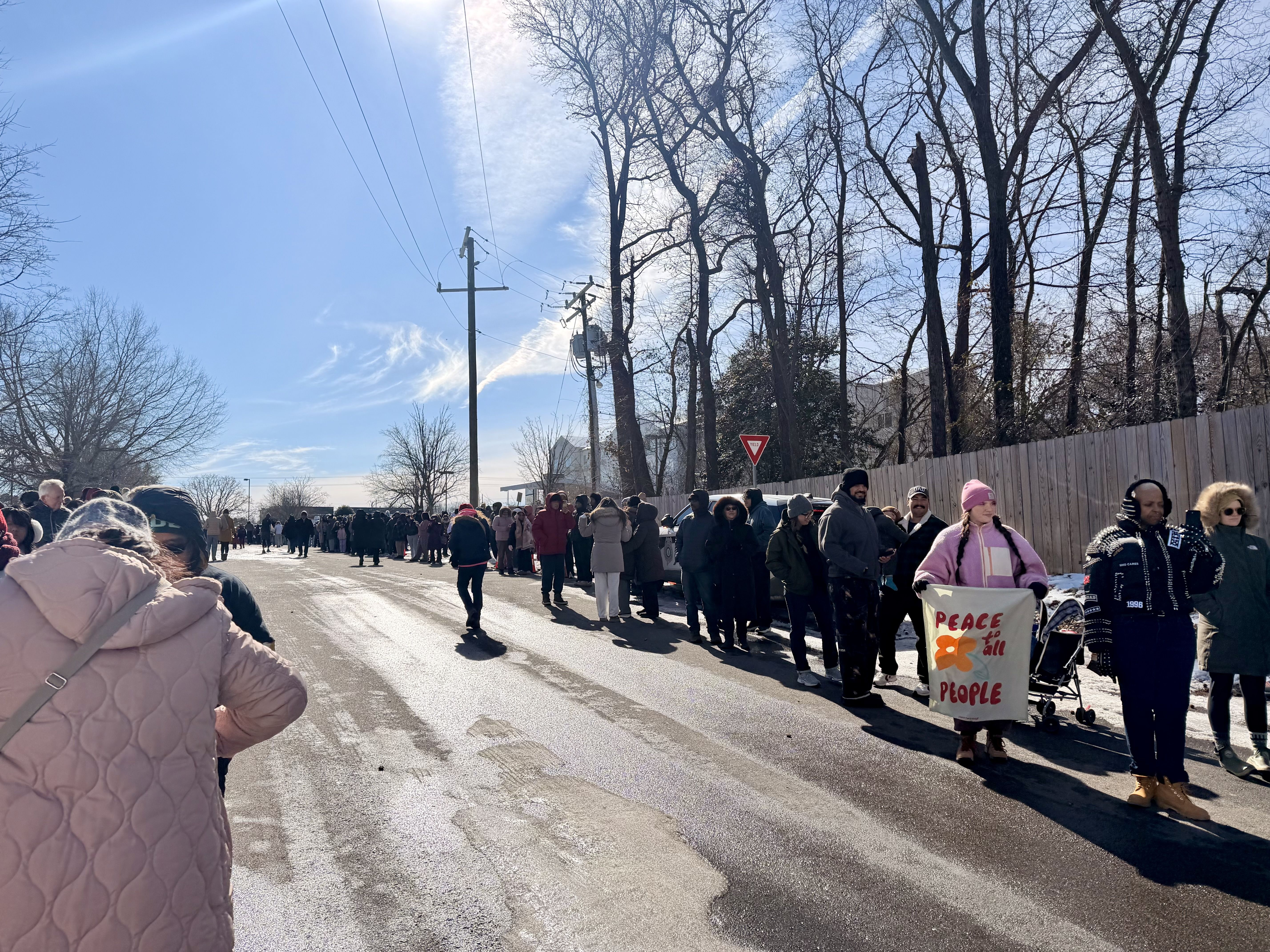 A diverse group of people stand in a long line along a sunny street with bare trees; one person holds a sign reading "Peace for all People" in red letters with an orange flower.