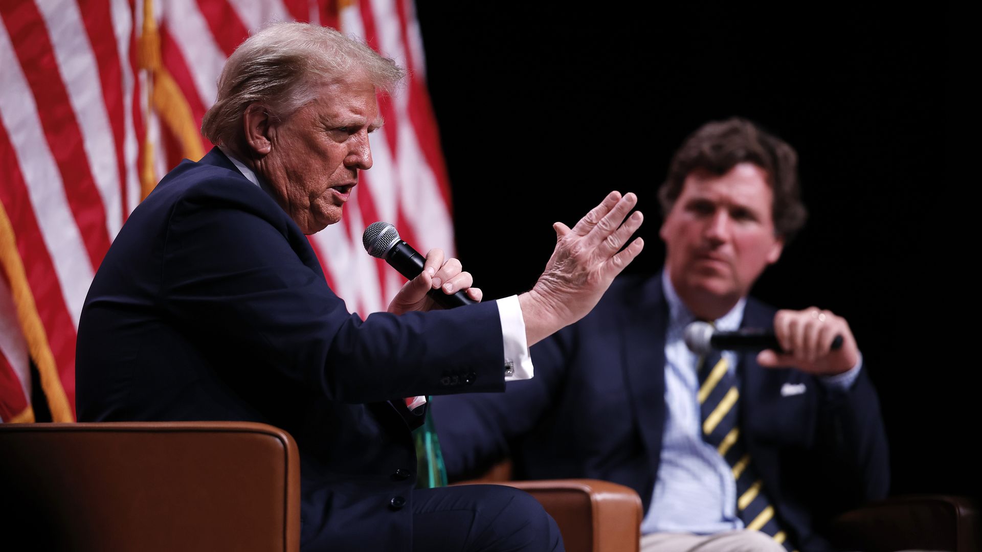 Trump, wearing a dark suit, gestures as he speaks into a microphone while sitting in a brown chair at an event. Tucker Carlson listens in the background.