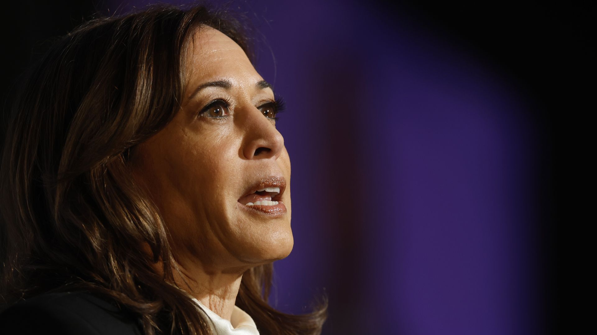 A close-up photo of Kamala Harris' face as she speaks with a purple background 