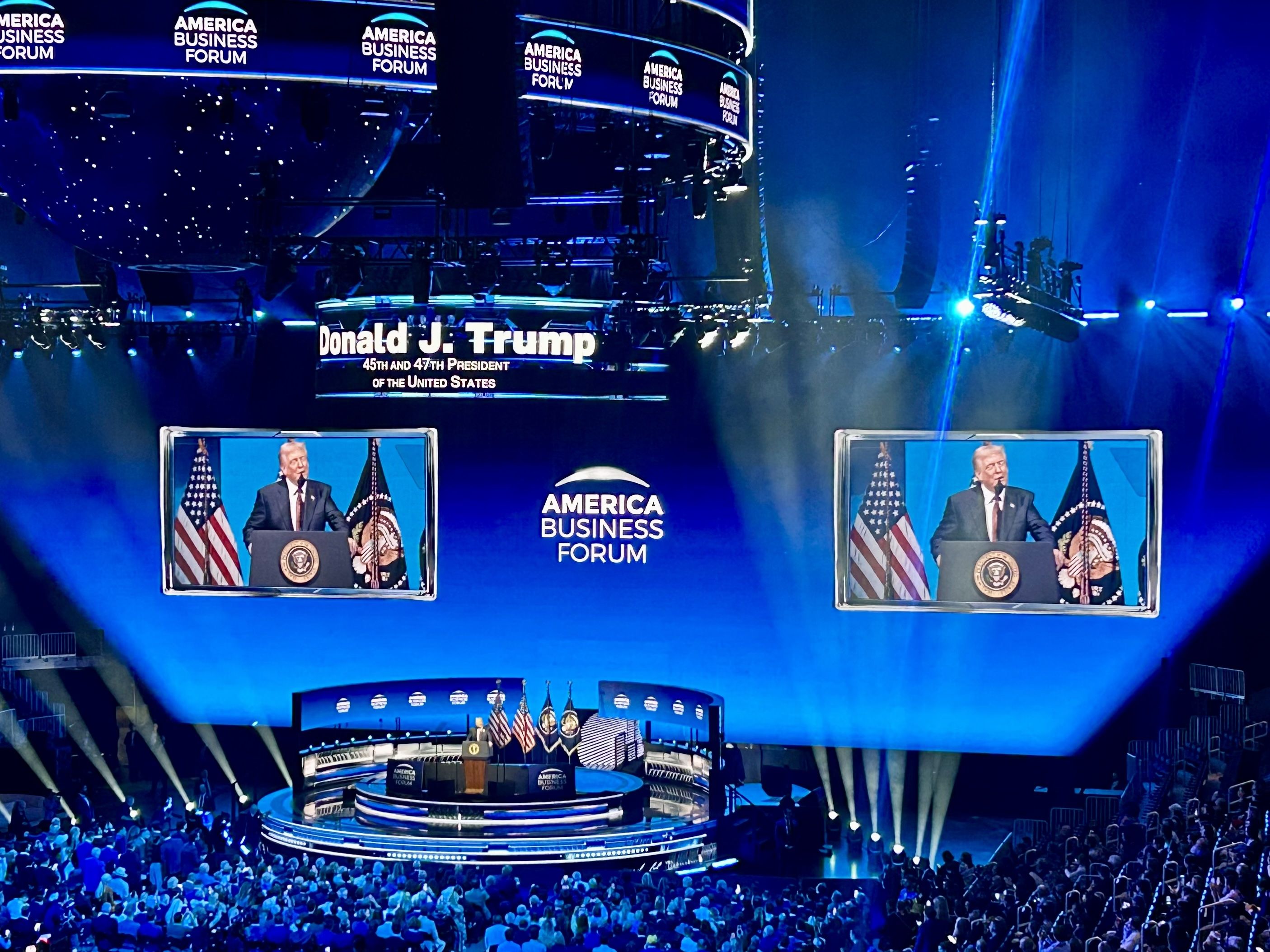 A large audience watches Donald J. Trump speaking at the America Business Forum on a circular stage with American flags, his image shown on two large screens above.
