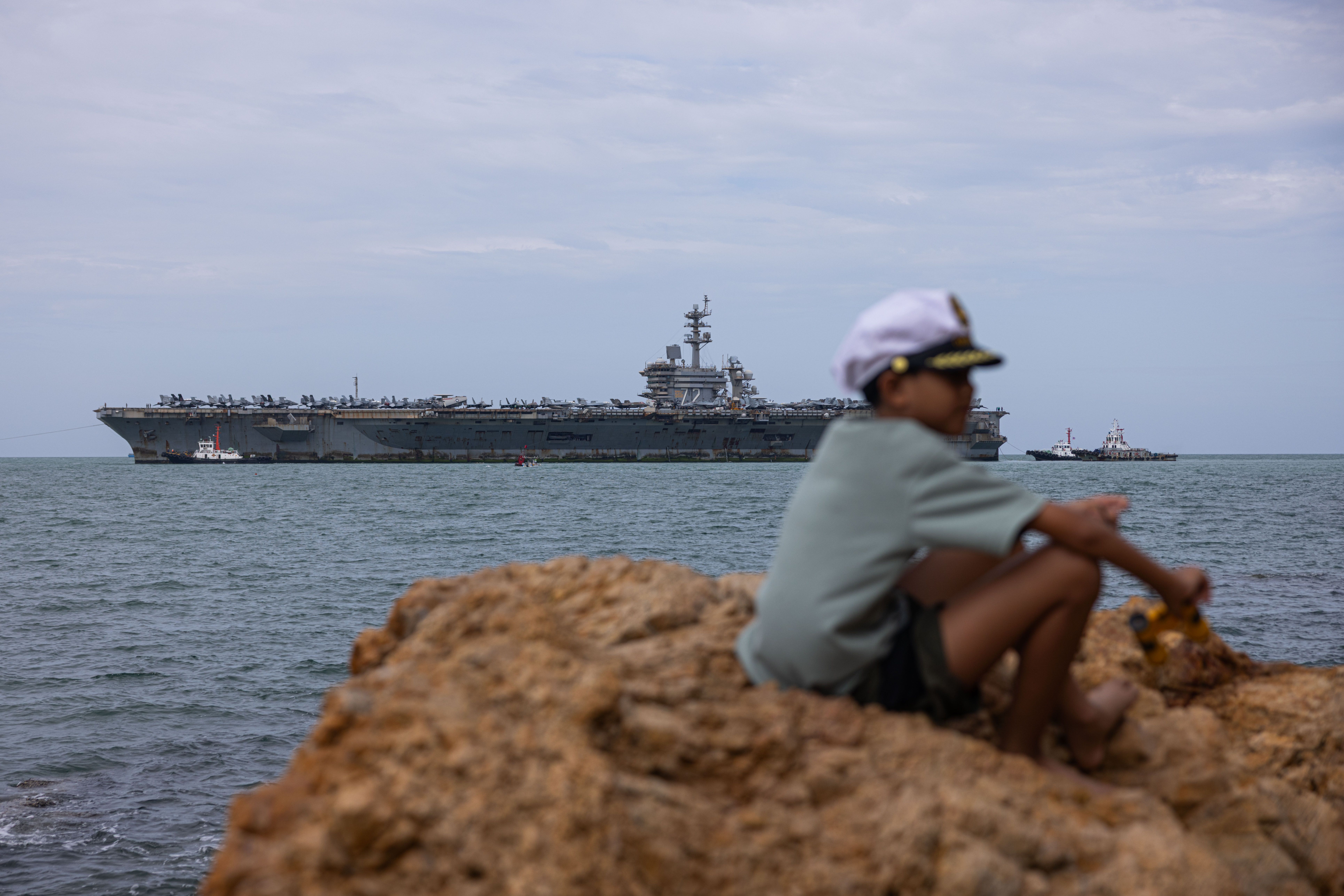 A young boy sits on a rock while wearing a sailor hat as the USS Abraham Lincoln, a large aircraft carrier, sails nearby.