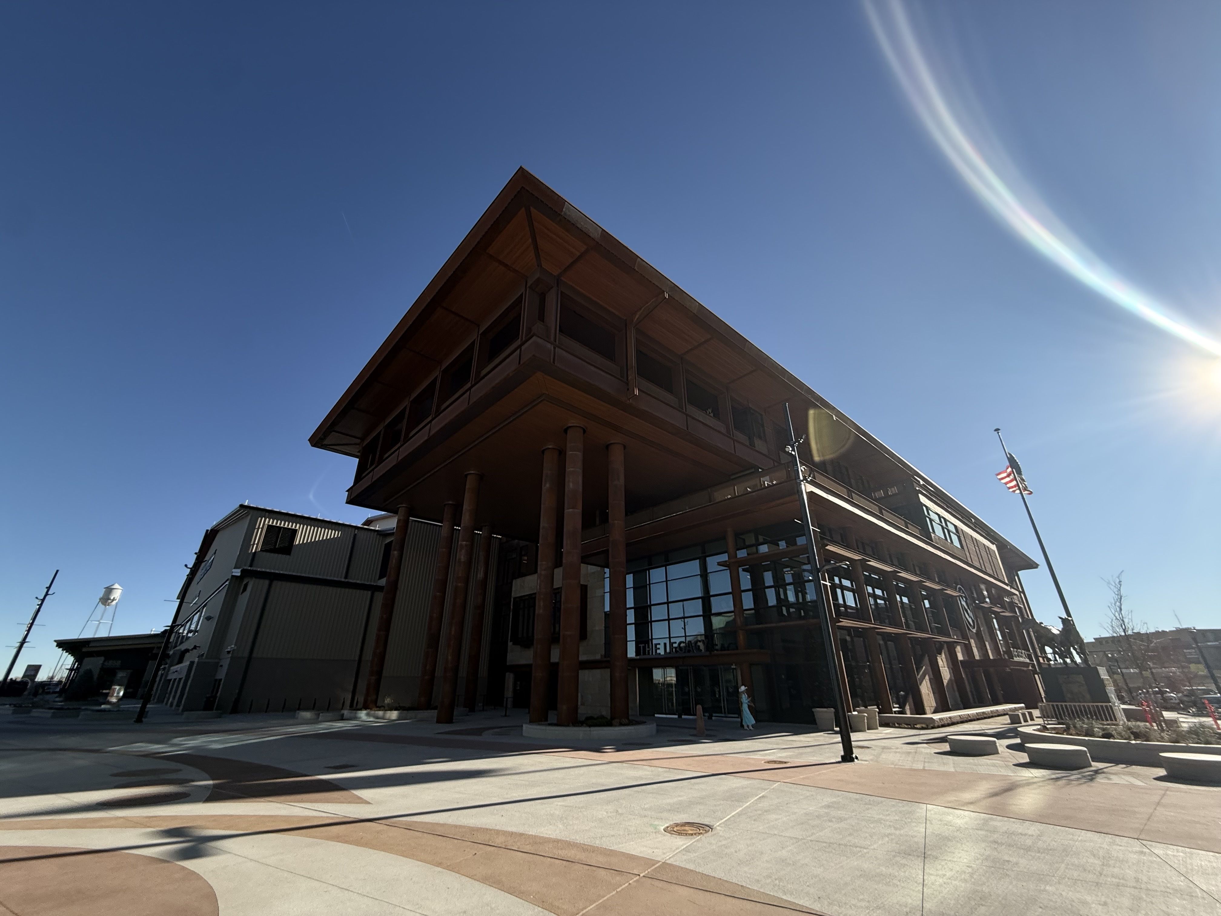 Modern building with tall rust-colored pillars and large glass windows under a clear blue sky, sunlight creating a lens flare, American flag on a pole, and paved open plaza.