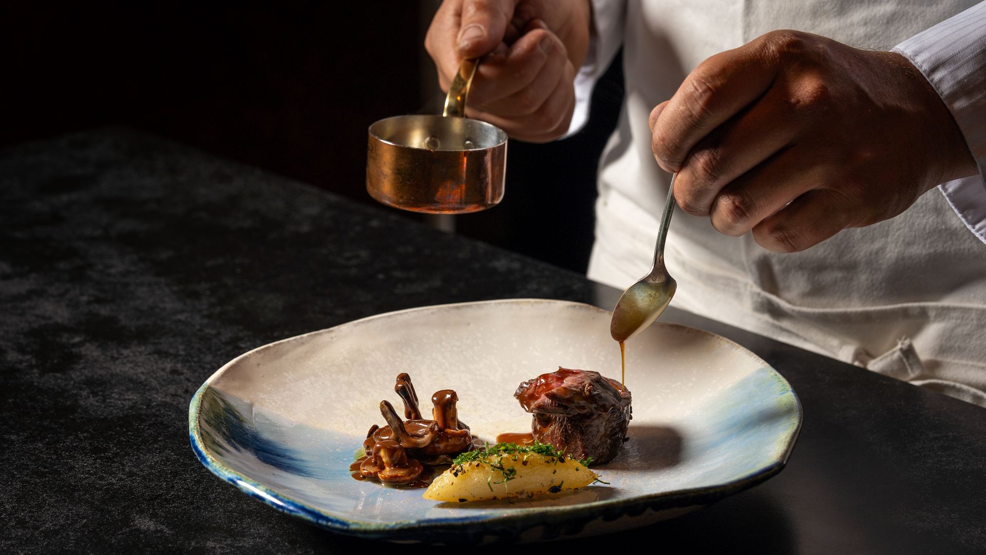 Close-up of a chef pouring warm brown sauce from a copper pot onto a plated dish. Hands hold the pot and spoon as sauce streams onto meat and vegetables.