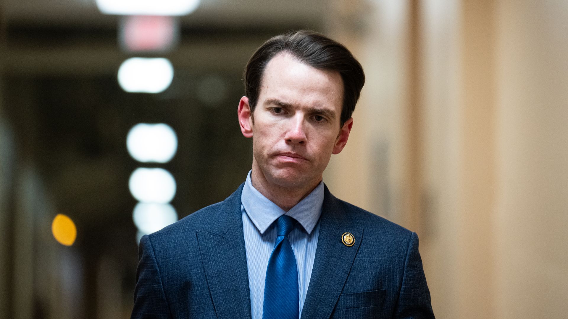 Man with dark hair, wearing a blue suit jacket, light blue shirt, and blue tie, standing in a hallway with blurred white and orange lights in the background.