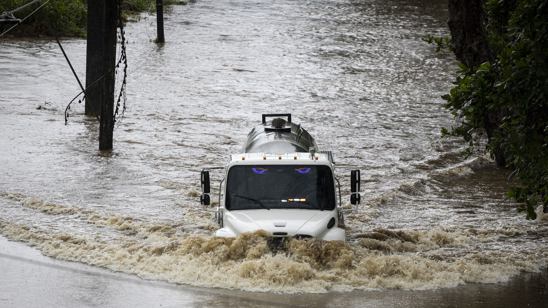 White tanker truck driving through a flooded muddy road with water splashing high, surrounded by trees and utility poles partially submerged in floodwater.