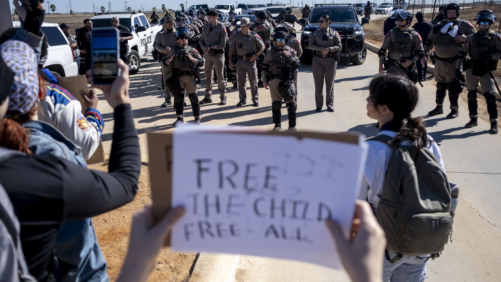 Protesters hold signs and phones while a large group of police officers in tactical gear stand on a sunlit road near police vehicles under a clear blue sky.