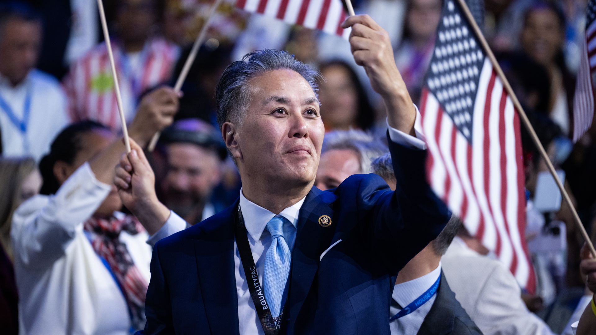 Rep. Mark Takano, wearing a blue suit and holding up an American flag while surrounded by a crowd.