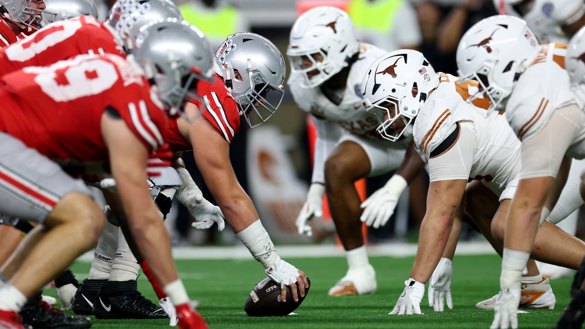 College football players from Ohio State in red and white helmets and Texas in white and orange helmets lined up face to face on the field, preparing for a play at the line of scrimmage.