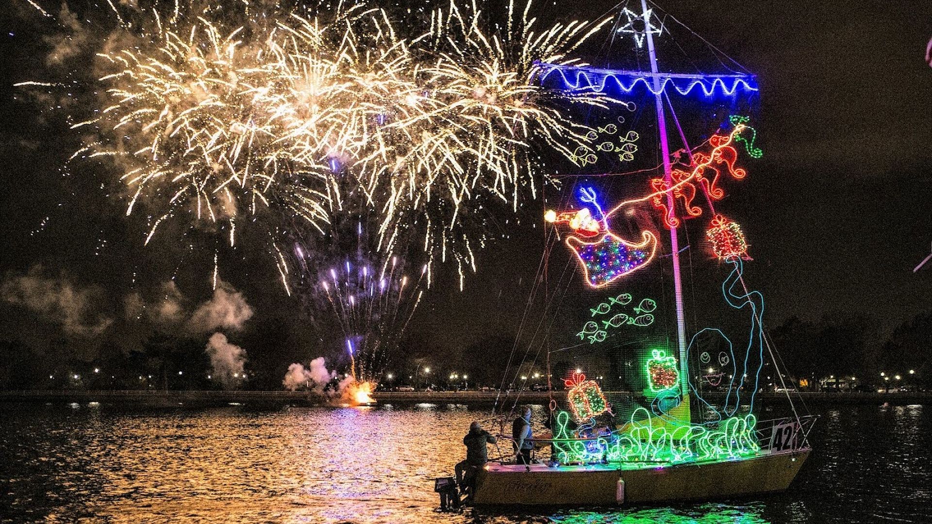 A small boat decorated with colorful Christmas lights on a dark lake at night, with fireworks illuminating the sky and reflecting on the water's surface.