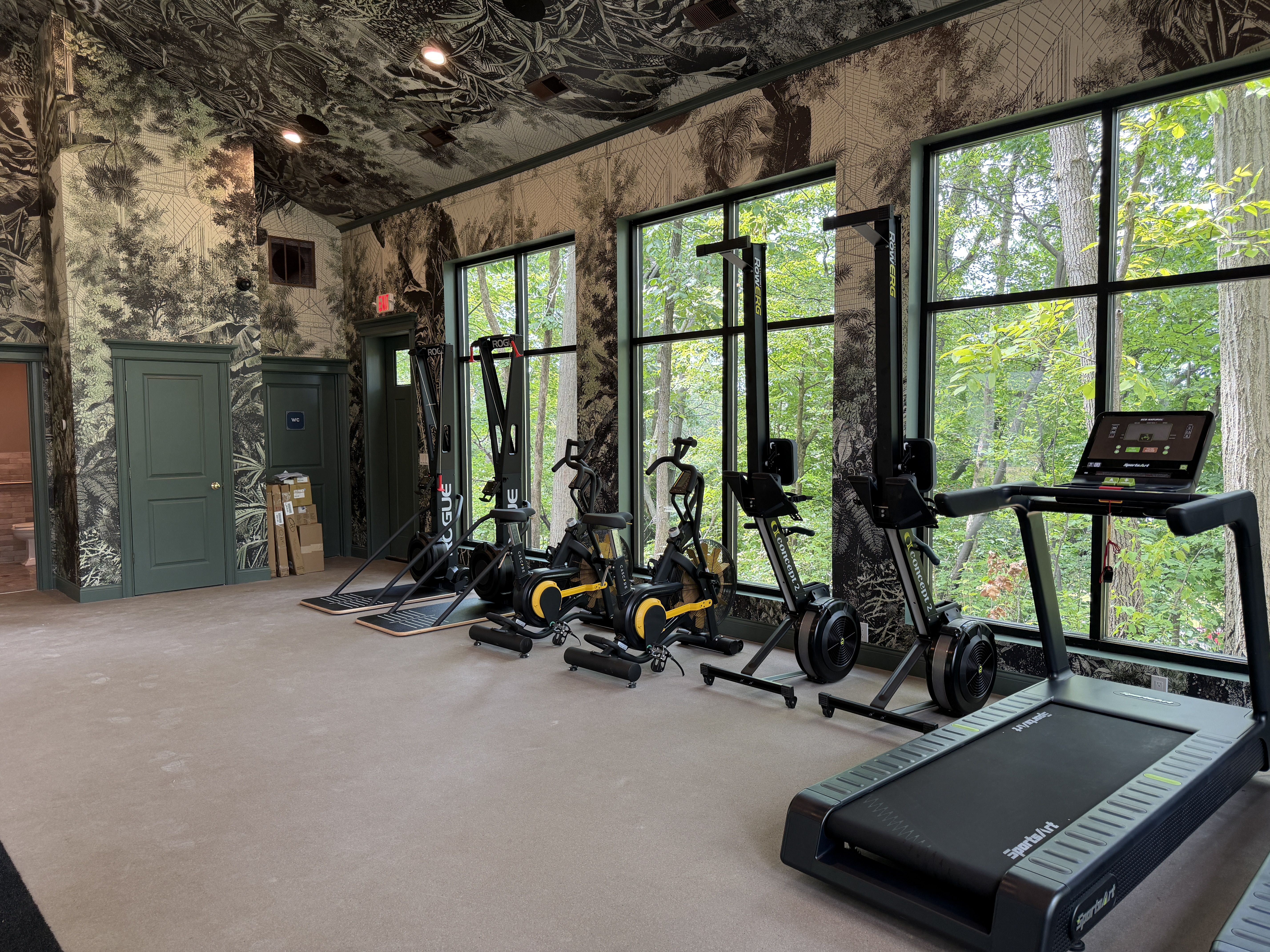 Indoor gym with a black-and-white botanical mural on walls and ceiling, a row of treadmills and bikes by large windows overlooking green trees outside; teal doors.
