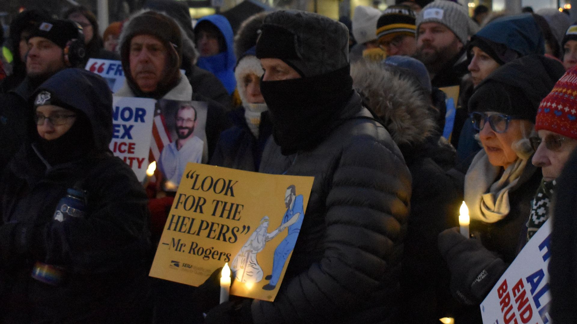 People in winter clothing holding candles and signs at a nighttime vigil, including one yellow sign saying \