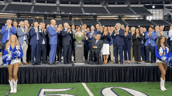 Group of formally dressed people on stage applauding around a trophy in a stadium with cheerleaders in blue and white outfits standing on grass marked with 50-yard line.