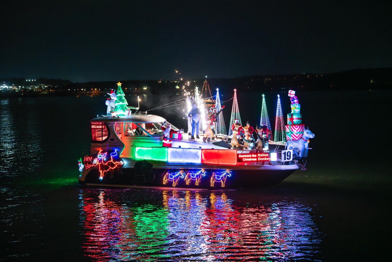 Boat decorated with colorful Christmas lights, inflatable Santa, Christmas tree, reindeer, presents, and characters illuminated at night, reflecting on water during holiday parade.