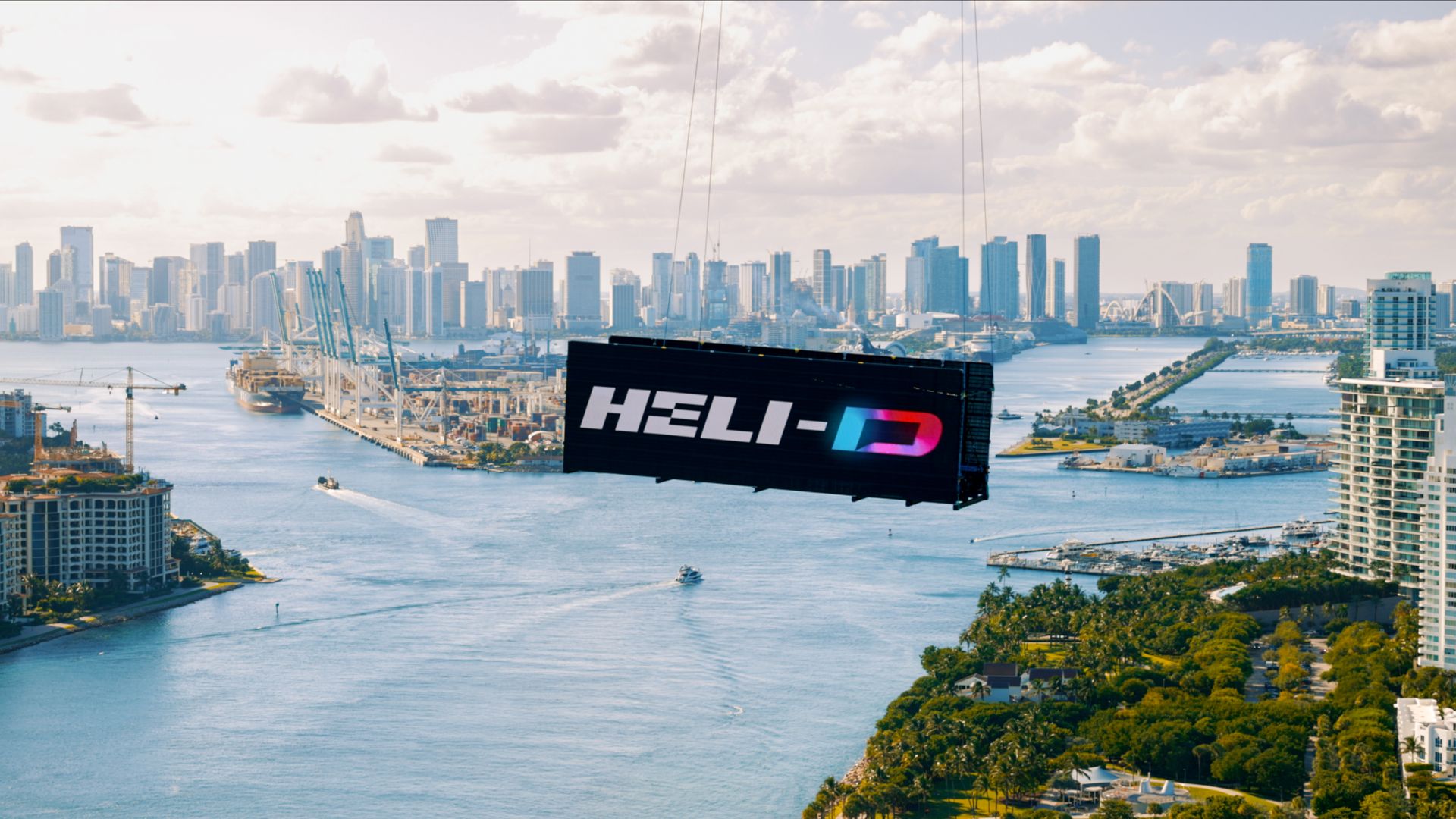 Large black billboard with "HELI-D" in white and gradient letters suspended over a waterway with boats, city skyline, and blue sky with clouds in the background.