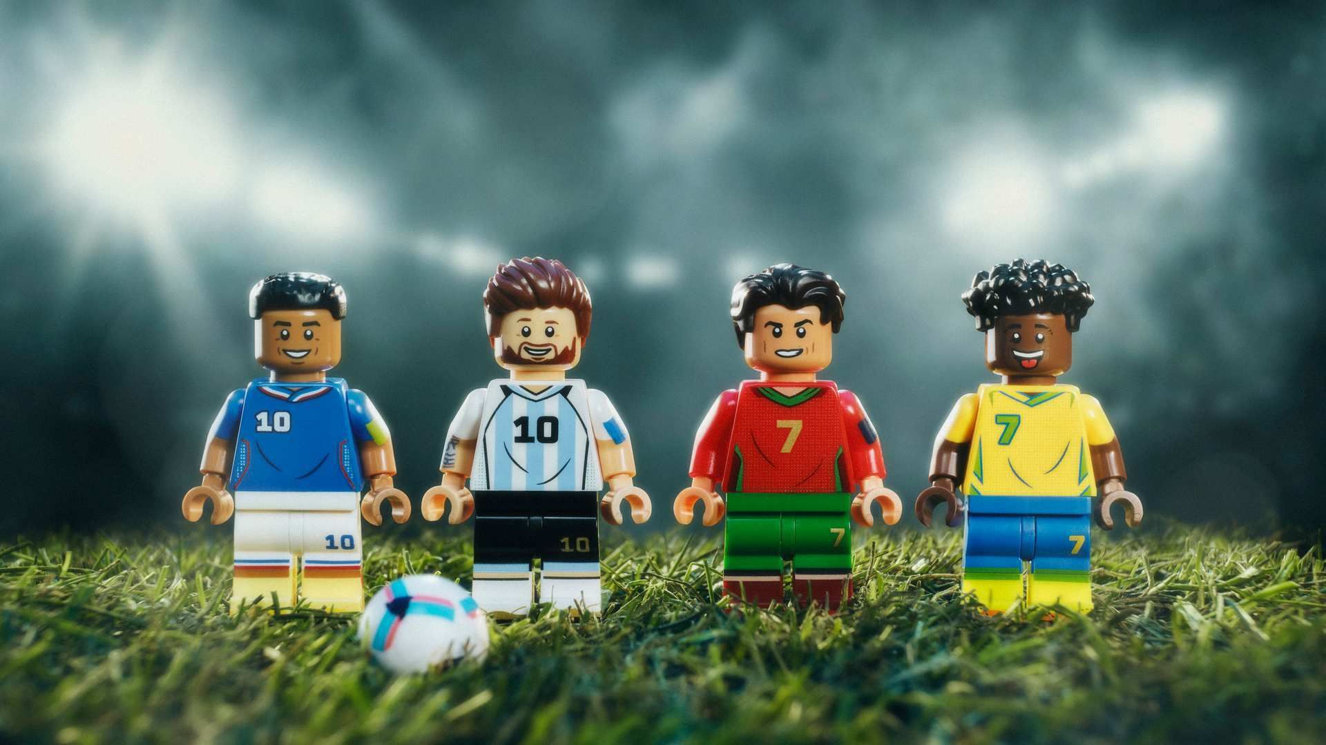 Four LEGO soccer players stand on grass, in blue, white, red, and yellow jerseys with numbers 10 and 7. A small ball rests near their feet as stadium lights blur in the background.