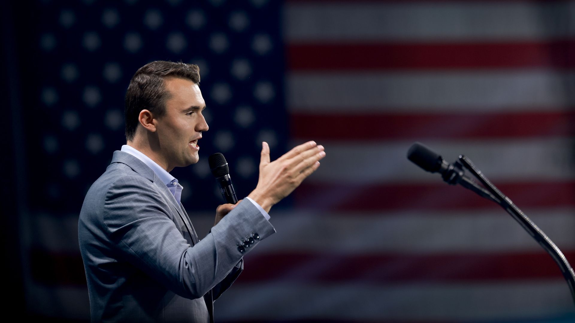 Charlie Kirk talking into a microphone with the American flag behind him