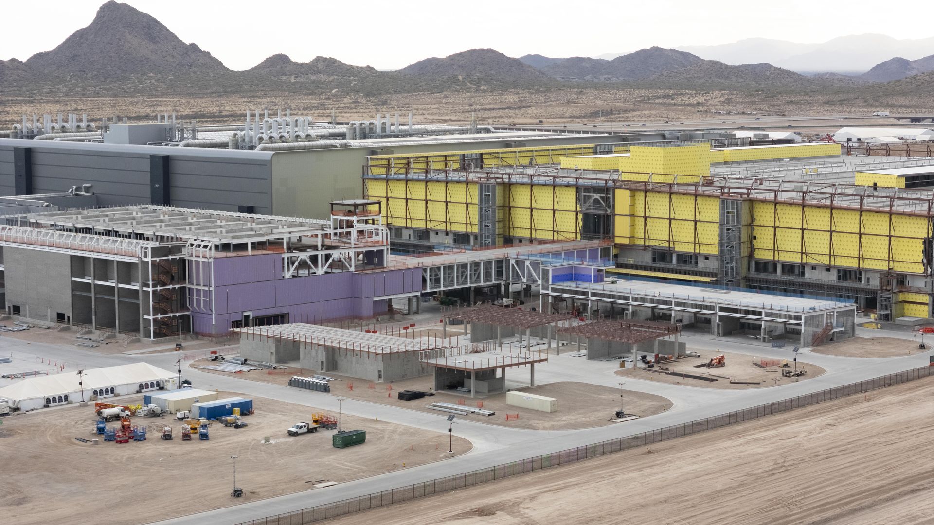 Large industrial facility under construction in a desert area with mountains in the background, featuring yellow and purple building panels and various construction equipment on site.