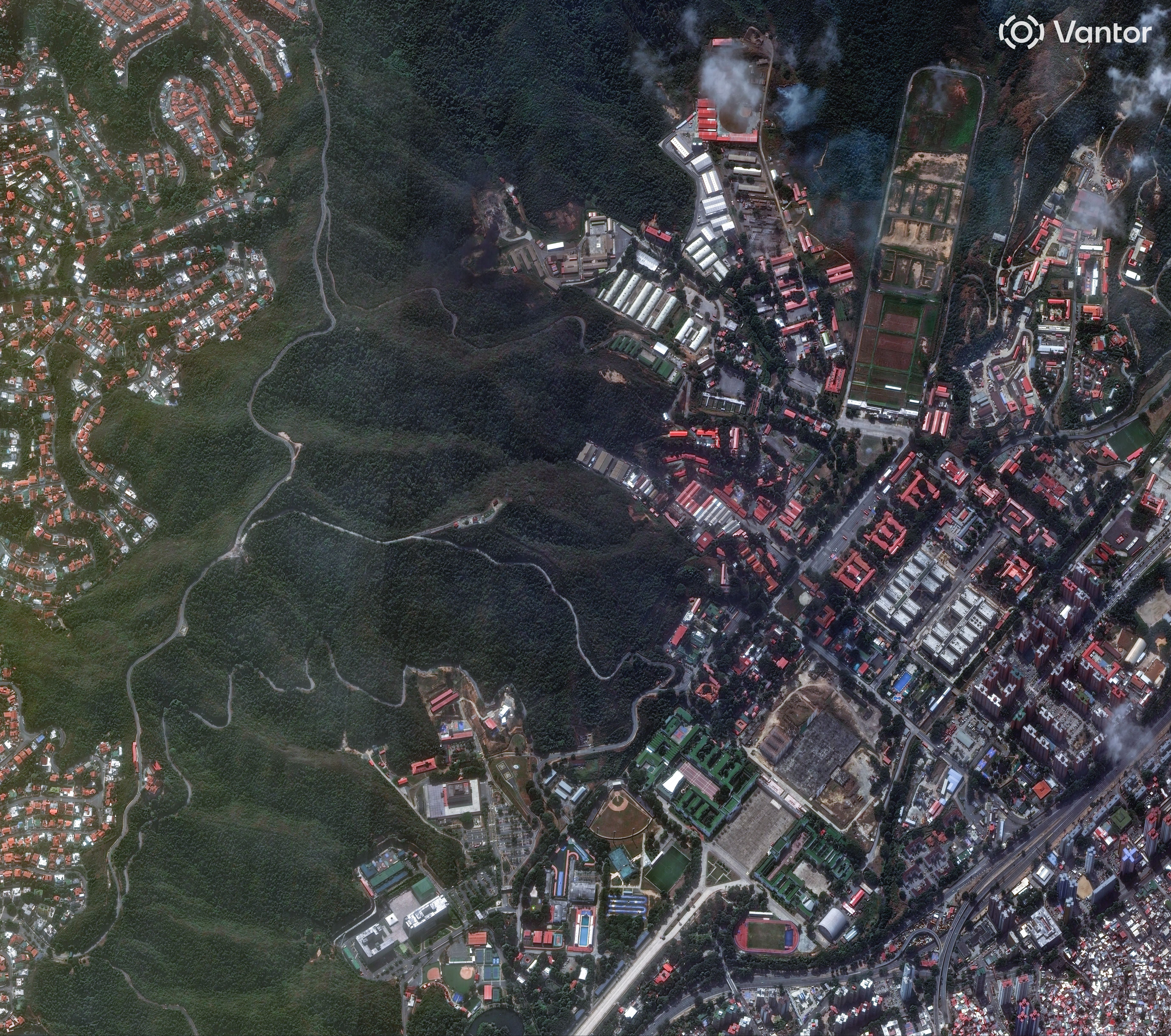 Satellite view of urban area with dense green forest in the center, roads winding through hills, clusters of red-roofed buildings, sports fields, and residential neighborhoods.
