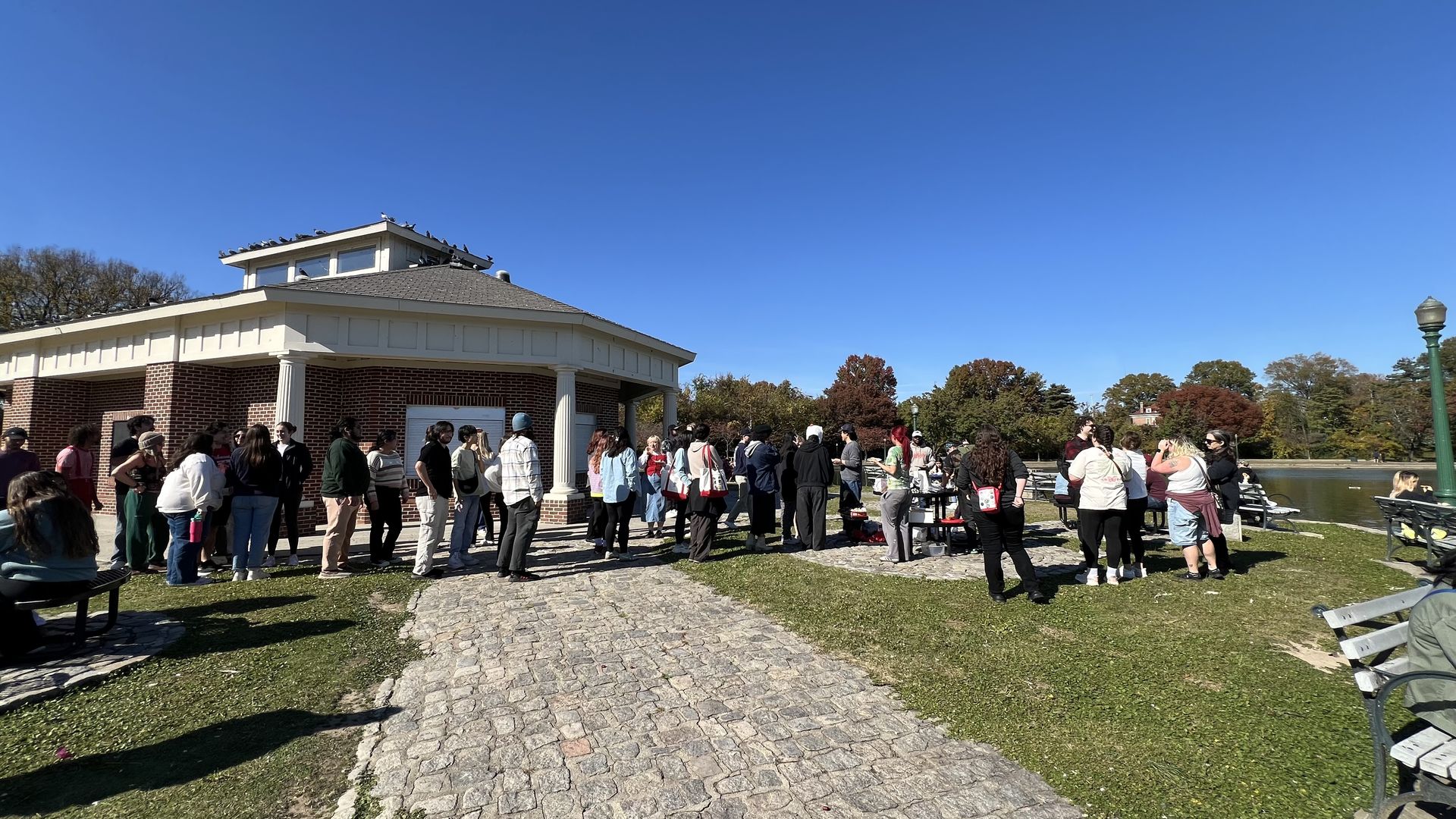 A group of people wait in line outside a small brick pavilion on a sunny day with a clear blue sky, near a pond and trees with autumn foliage.