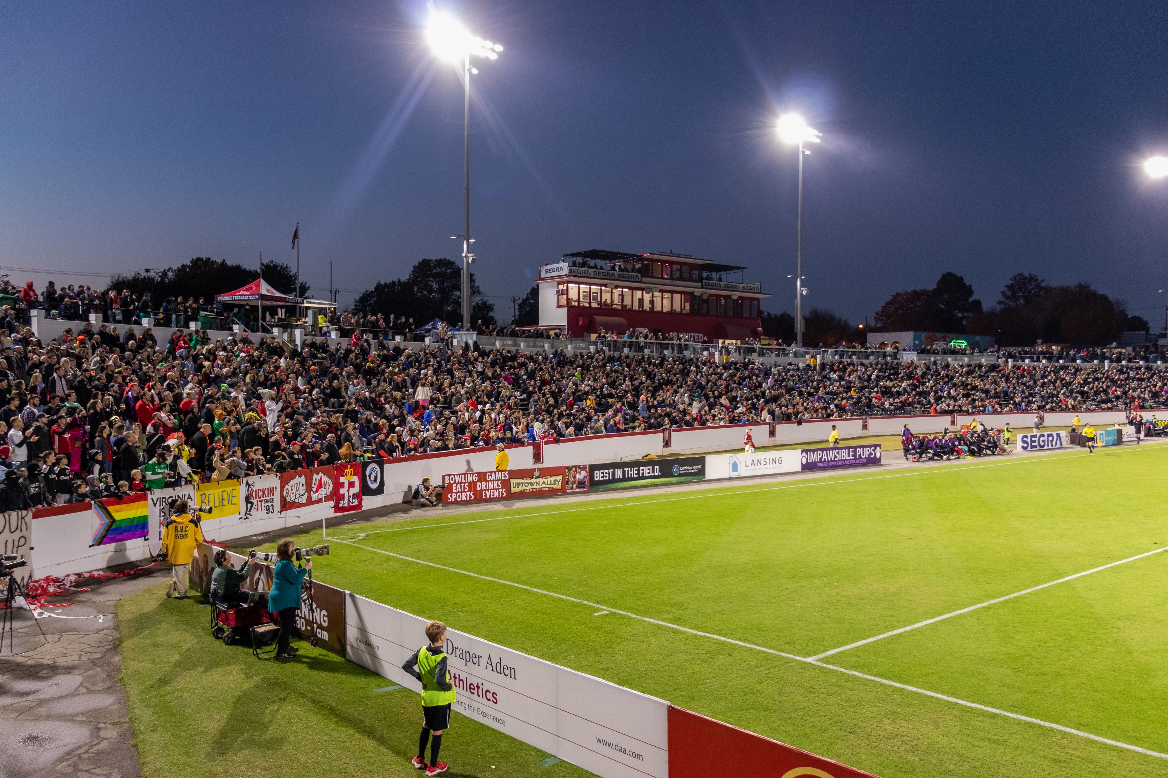 Nighttime stadium scene with a packed stands and colorful banners along the wall. Bright floodlights cast glare over a red, two-story press box as spectators watch a game on the green field.