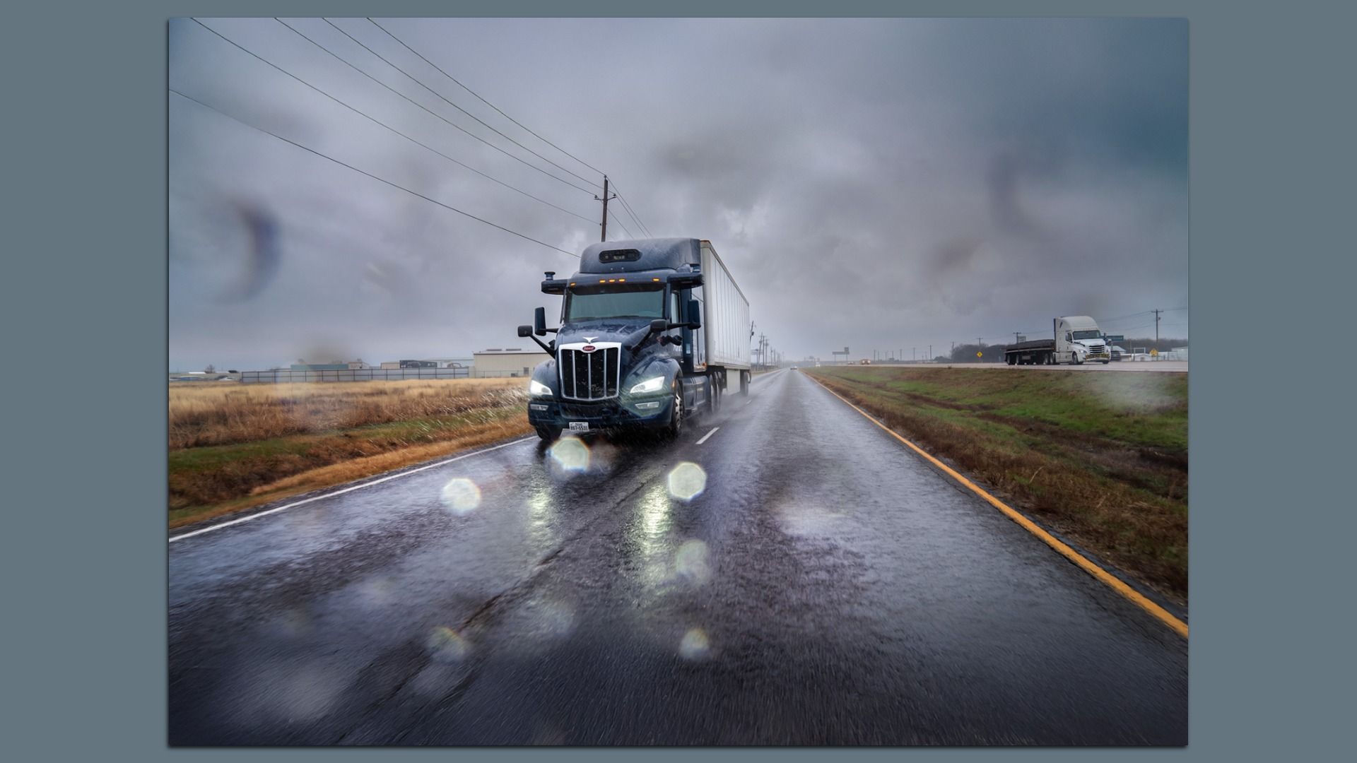 A driverless black semi-truck with headlights on drives on a wet road under gray, rainy cloudy sky with raindrops on the camera lens and dry grassy fields on both sides.