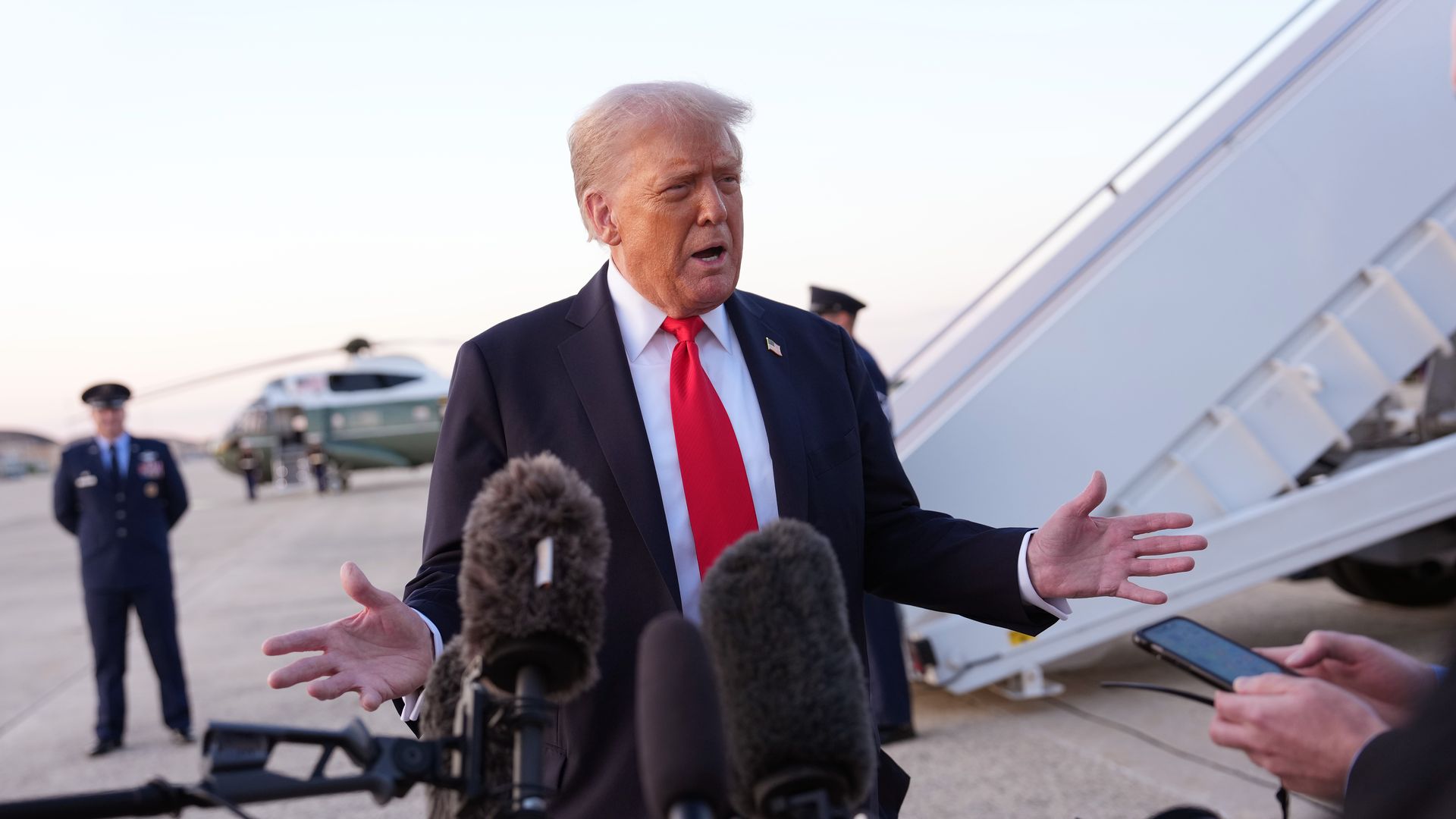 President Trump, wearing a black jacket with US flag pin, white shirt and red tie, speaks to reporters in front of four large mics at  Joint Base Andrews, Maryland,  as a military official in navy uniform and many medals looks on in front of a green helicopter.