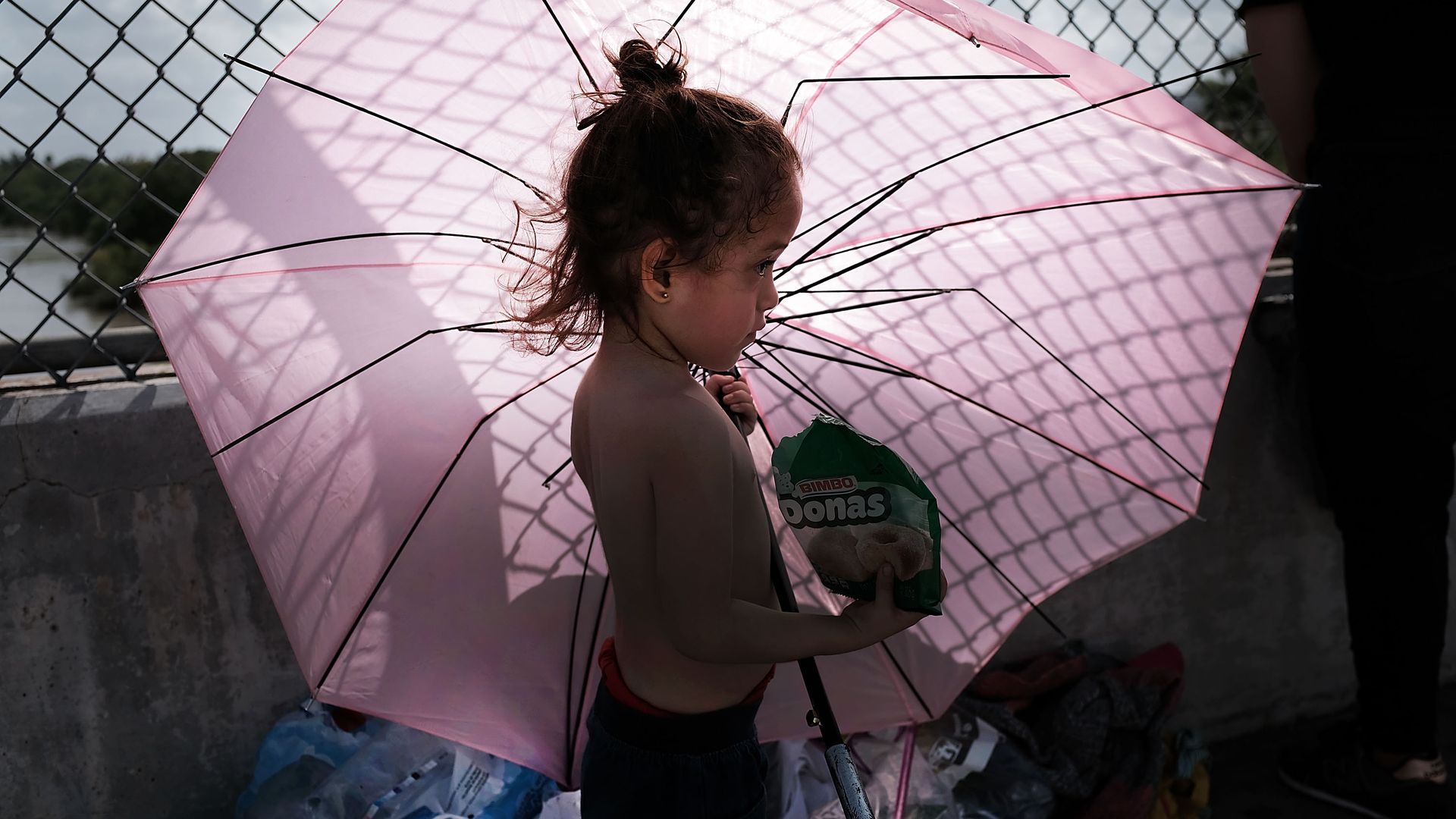 A child migrant holding a bag of chips and a big, pink umbrella