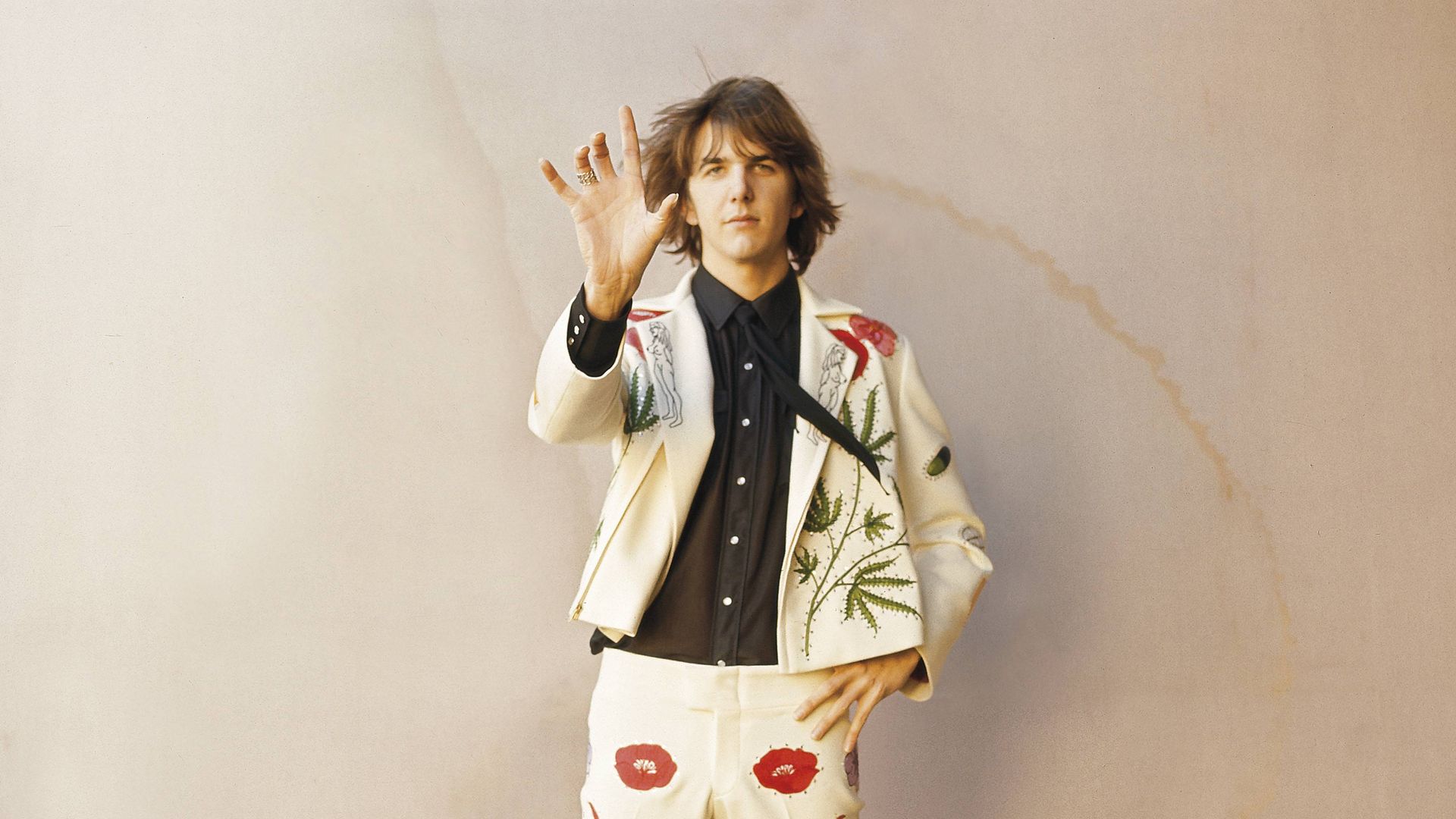 Man with shoulder-length hair wearing a cream suit with red patterns, black shirt, standing on a wooden platform and raising his hand toward the camera against a plain beige wall.