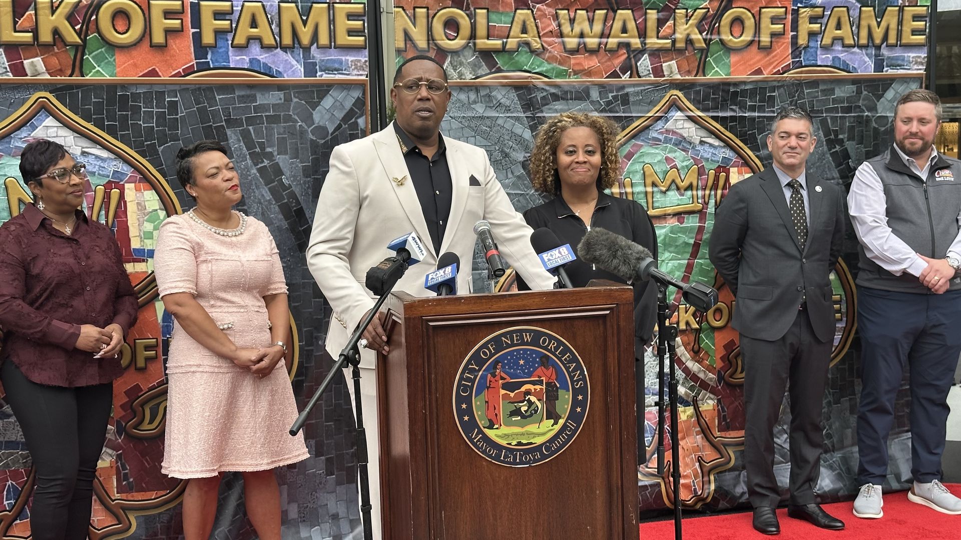 Master P speaks at a podium with the City of New Orleans' seal next to Mayor LaToya Cantrell, Syrita Steib and other leaders.