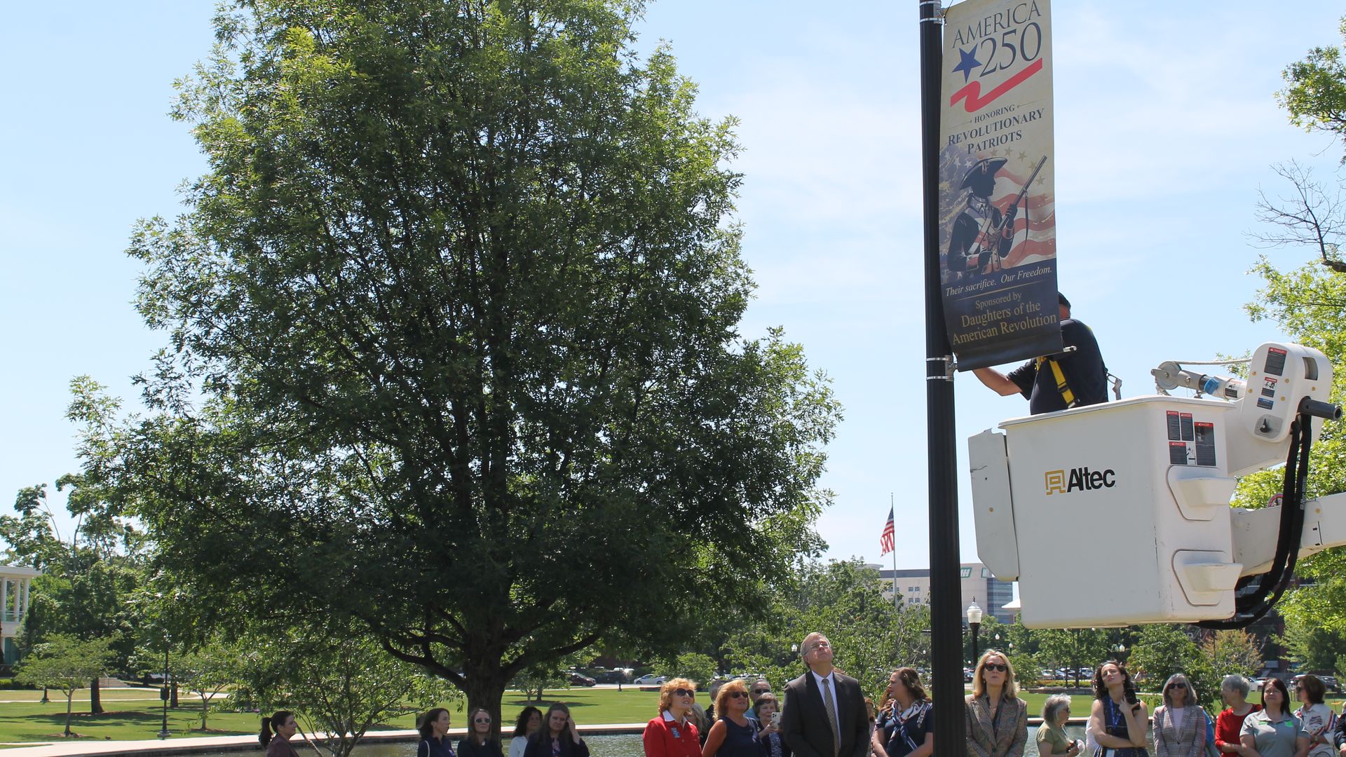 Crowd by a pond watches a tall lamppost banner reading America 250 with a star and flag; a worker on a white Altec bucket truck stands beside a large tree under a blue sky.