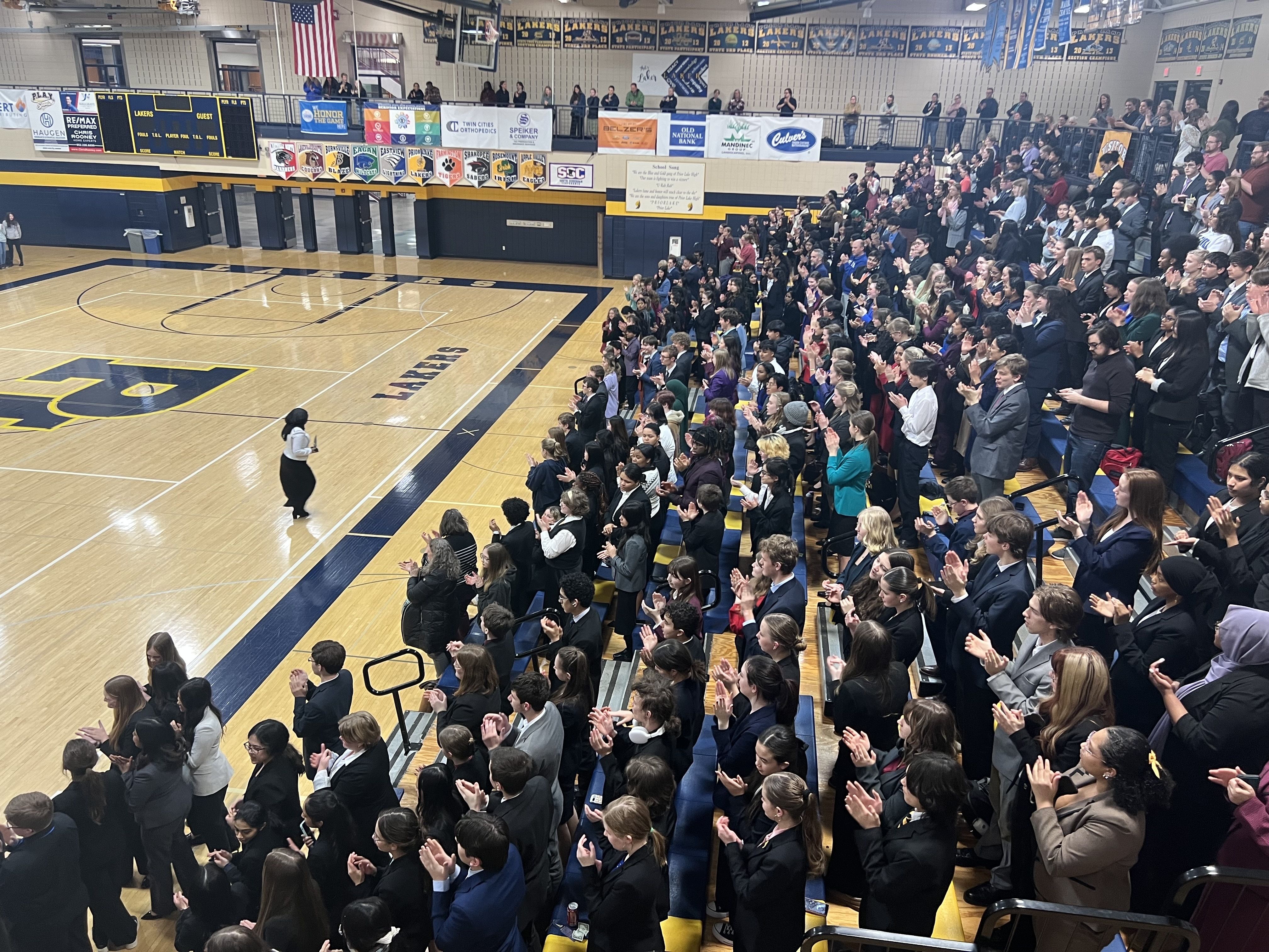 A high school gymnasium crowd stands and applauds as an award-winner strides across the basketball court