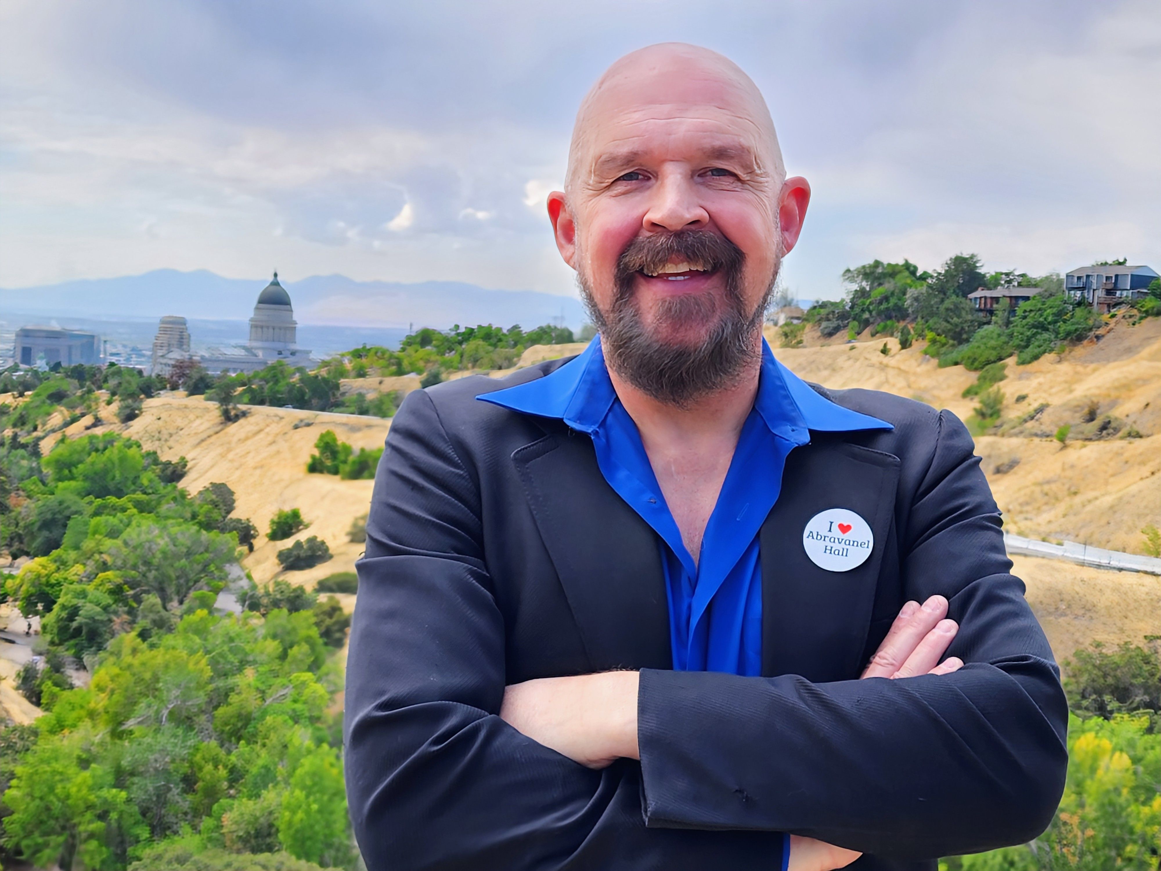 Smiling bald man with a beard wearing a blue shirt and black blazer with "I ♥ Abravanel Hall" button, standing outdoors with green trees and a domed building in the background.