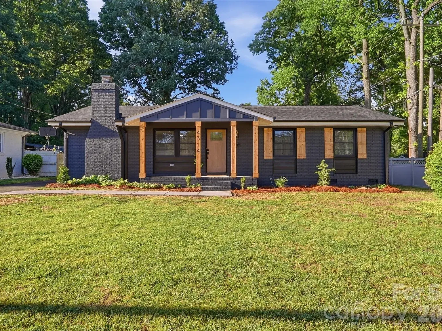 Single-story dark brick house with a front porch supported by light wood columns, a stone chimney on the left, triangular glass gable, black-framed windows, and a well-kept lawn with trees in the background.