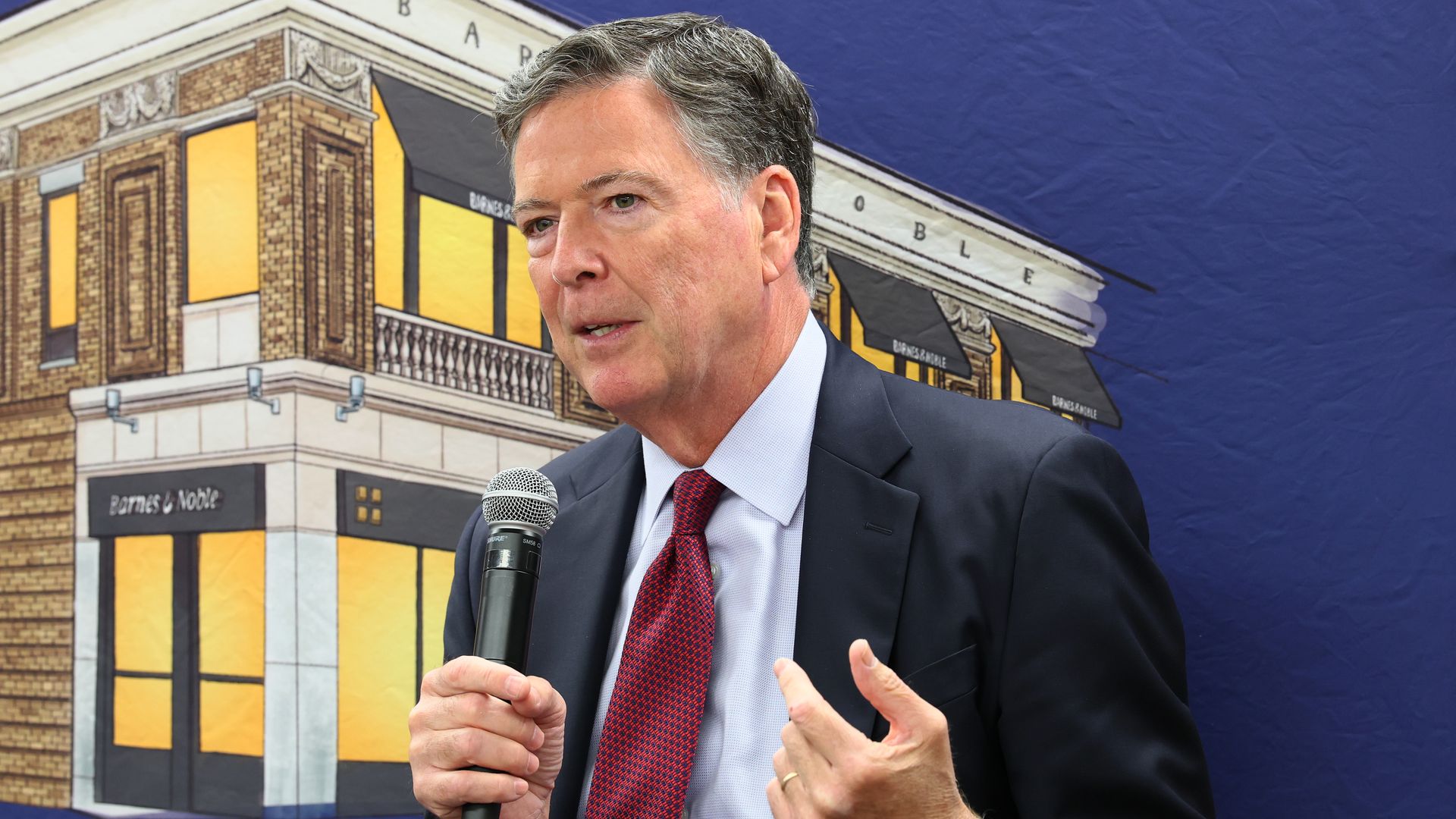 A graying, black-haired former FBI Director James Comey gestures with his left hand as he holds a mic in his right hand in front of a mockup of a building with lights on and a blue screen that signifies night time.