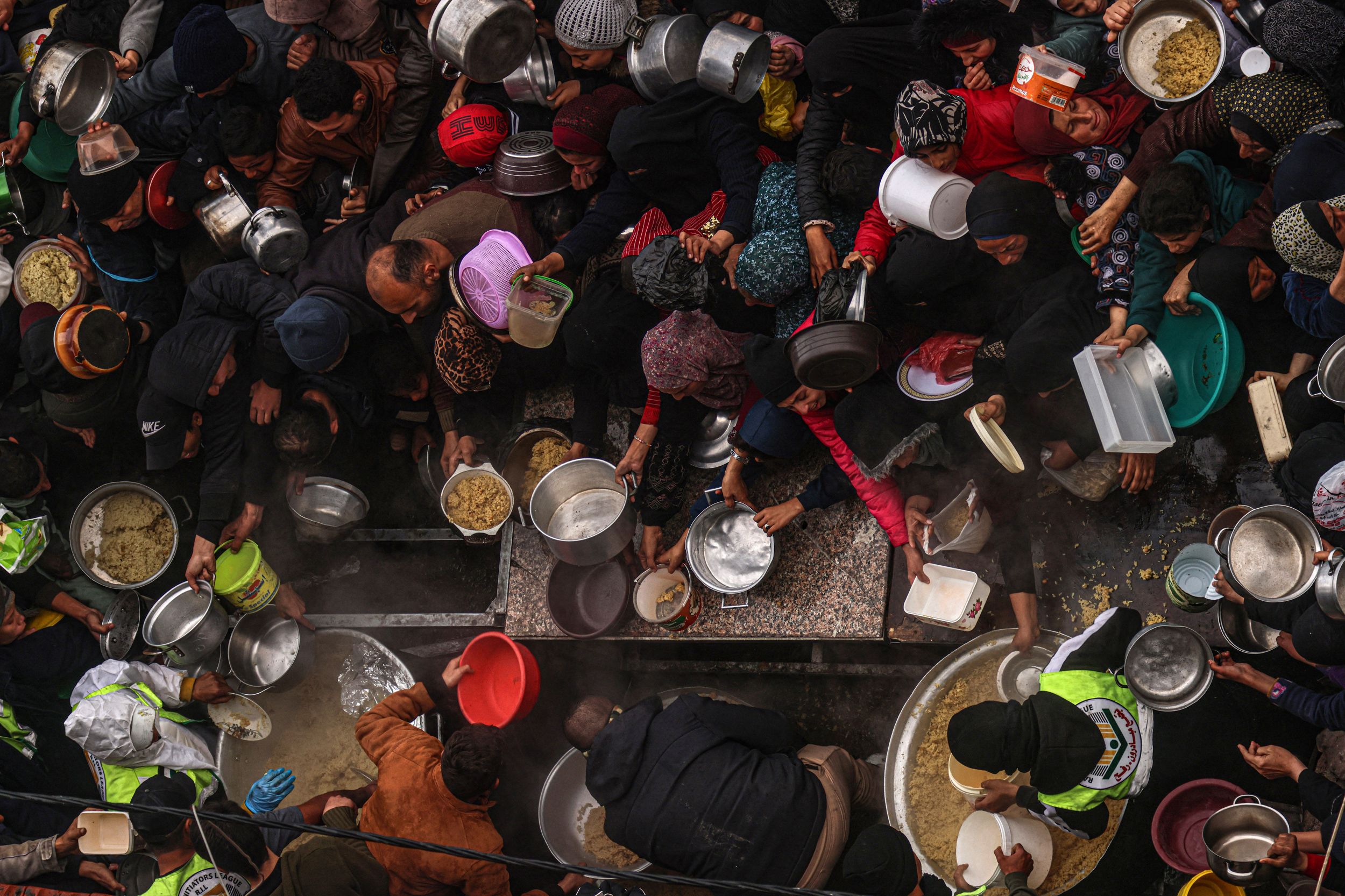 A large group of people holding various pots and pans, reaching out and pushing for food.