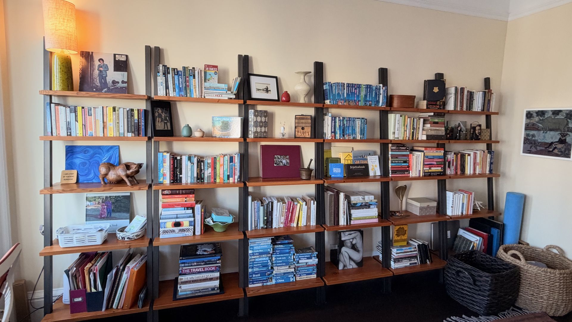 A tall floor lamp glows beside a wall of orange-brown shelves filled with books, framed photos, vases, and small decor. Beige walls with crown molding; a large woven basket on a dark rug.