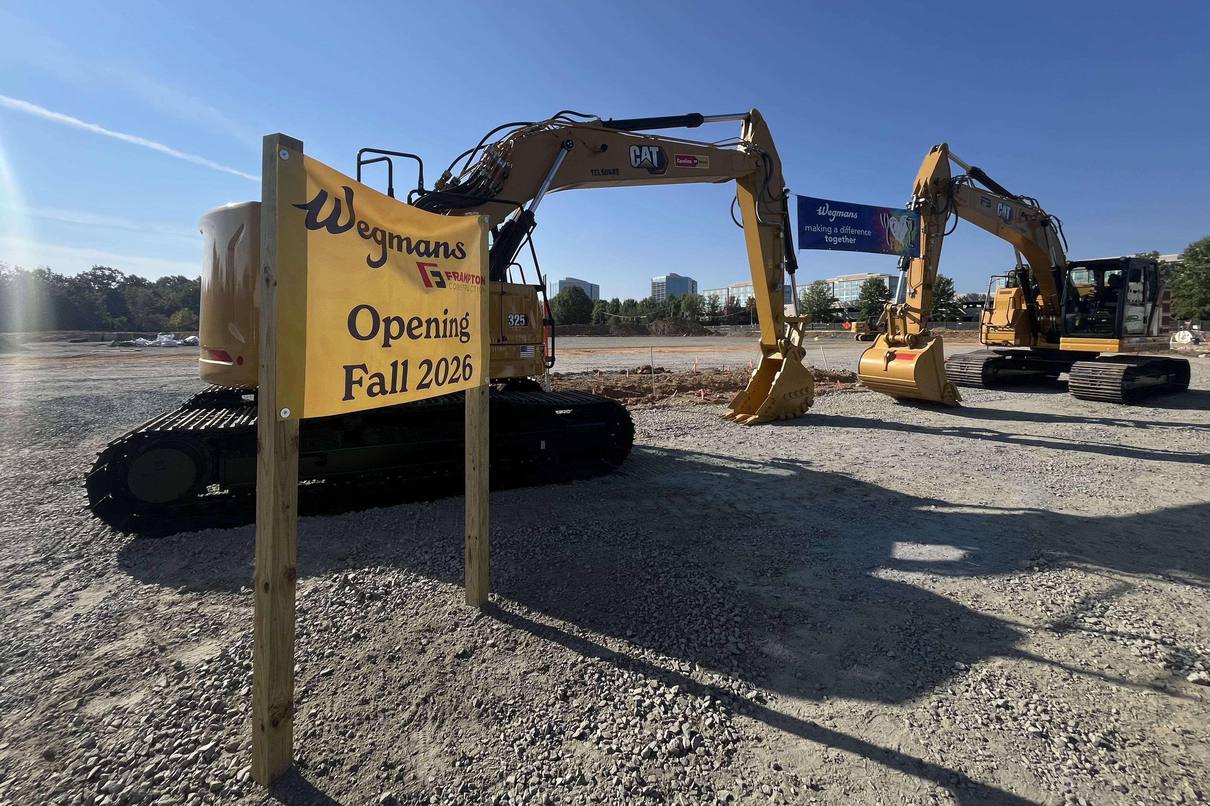 Construction site with two yellow CAT excavators and a yellow sign announcing Wegmans opening Fall 2026, under a clear blue sky with some buildings in the background.