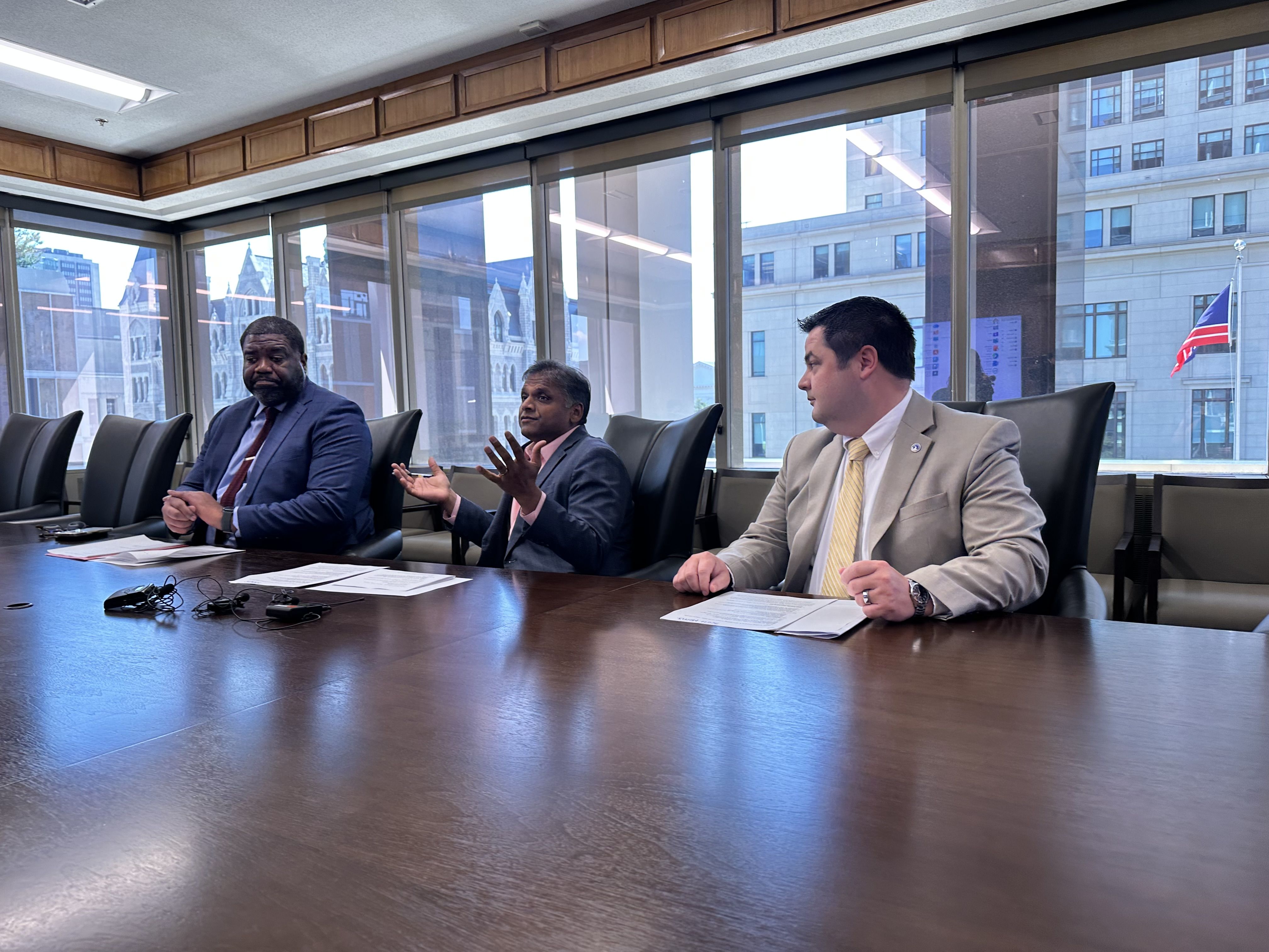Three men in business suits sit at a long conference table in a glass-walled office; papers and microphones lie on the table as city buildings and a Norwegian flag appear outside.