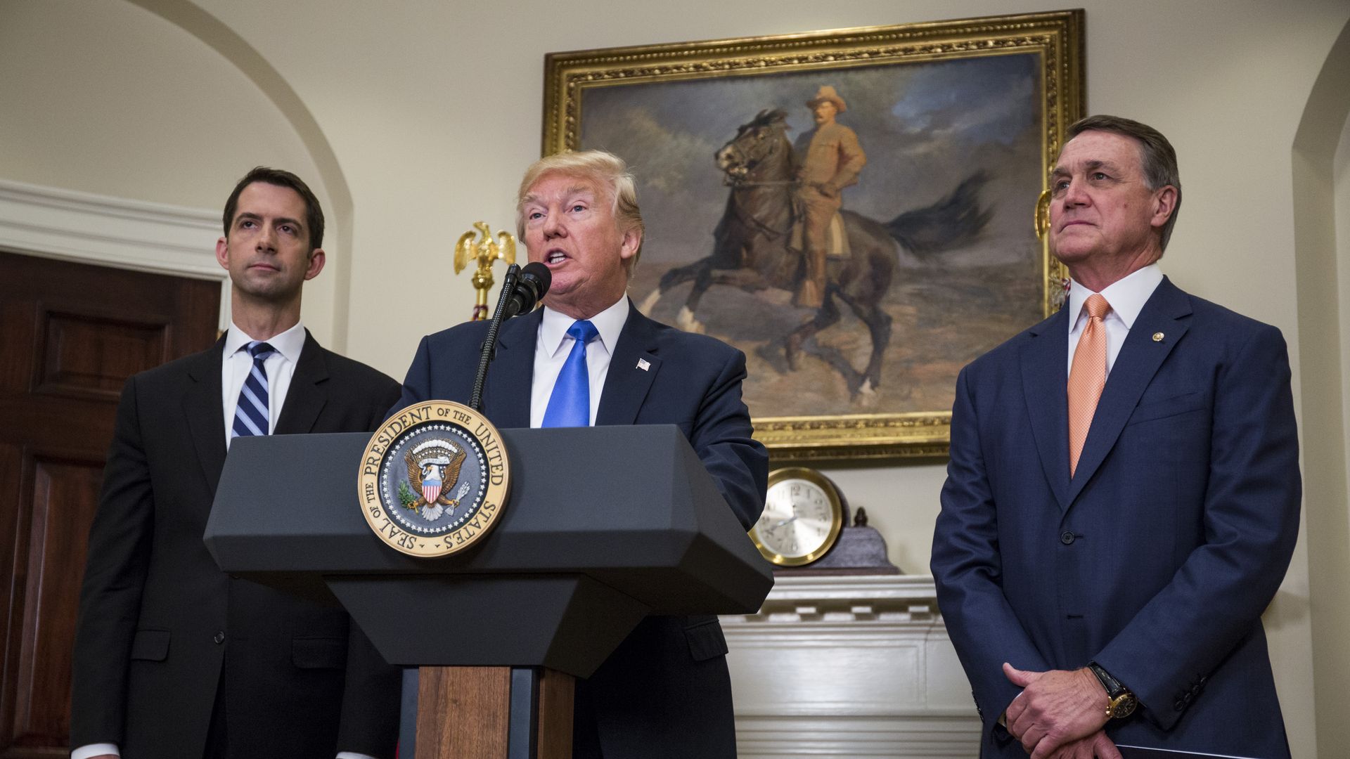 Photo of Donald Trump speaking from a podium as David Perdue stands next to him with his hands crossed in front