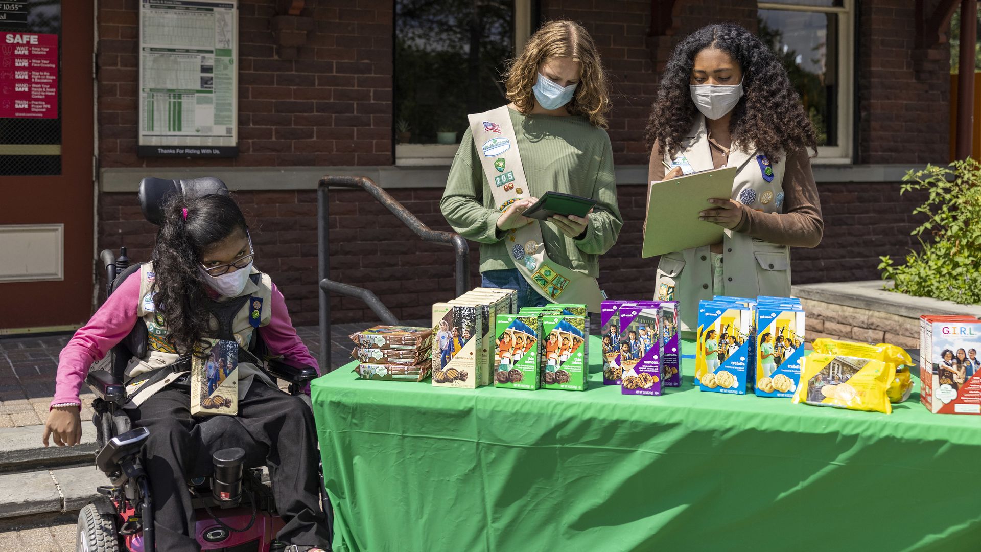Two girls in beige vests and one in a wheelchair sell Girl Scout Cookies at a green-clothed table outdoors.