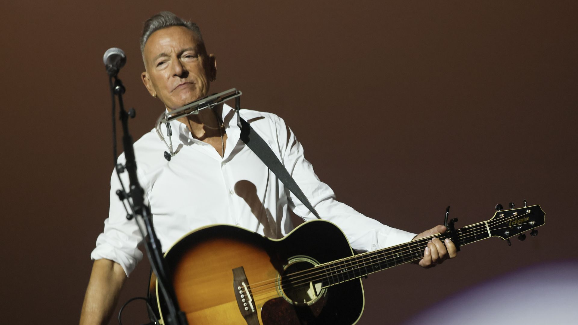 Bruce Springseen wearing a white shirt holds a brown guitar and has a harmonica around his neck. He's in front of a microphone. 